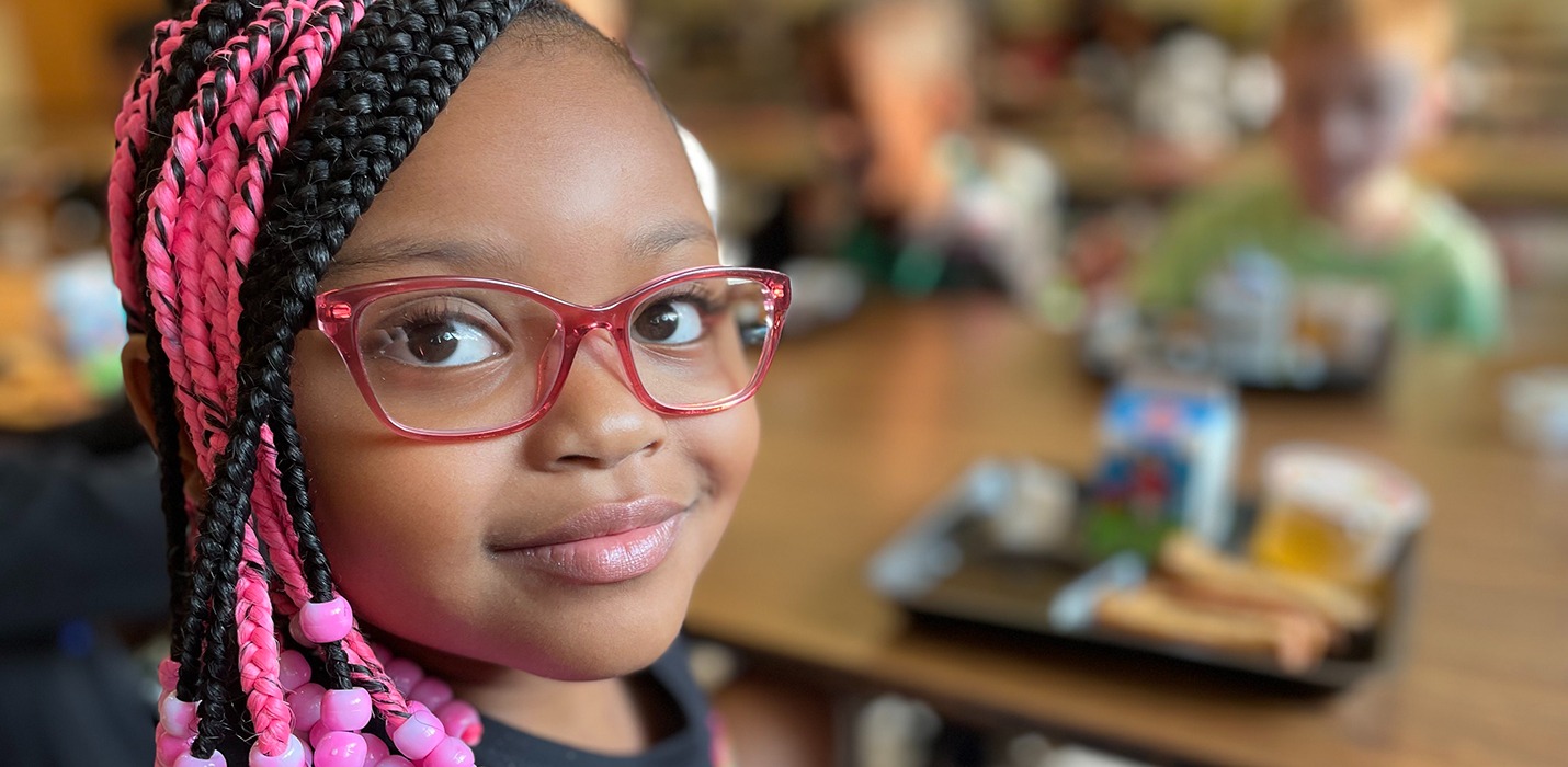 Elementary-age girl smiling looking at the camera. Her lunch tray with food is blurred in the background along with other students eating lunch.