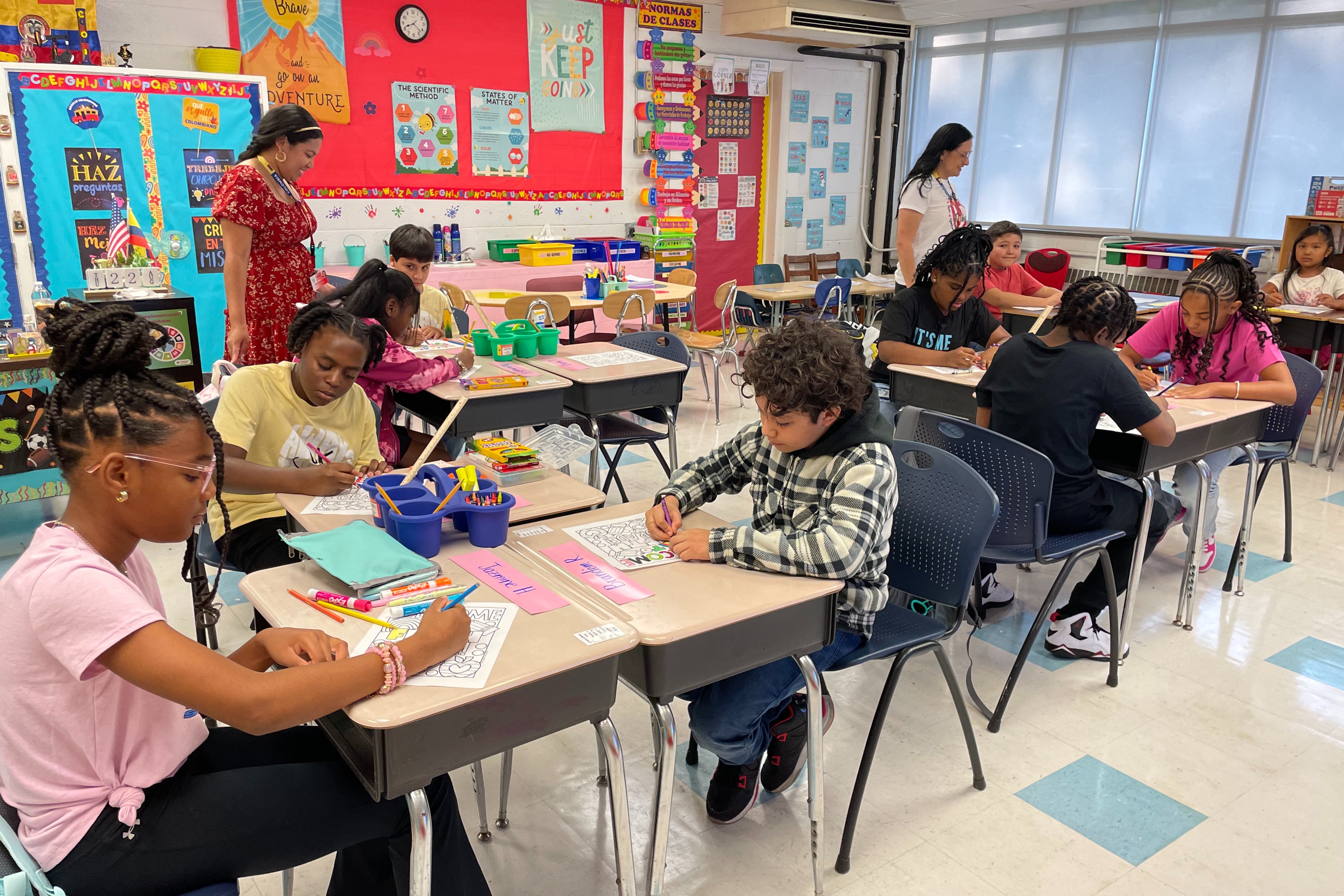 students working at their desks in a classroom with teachers smiling and watching them