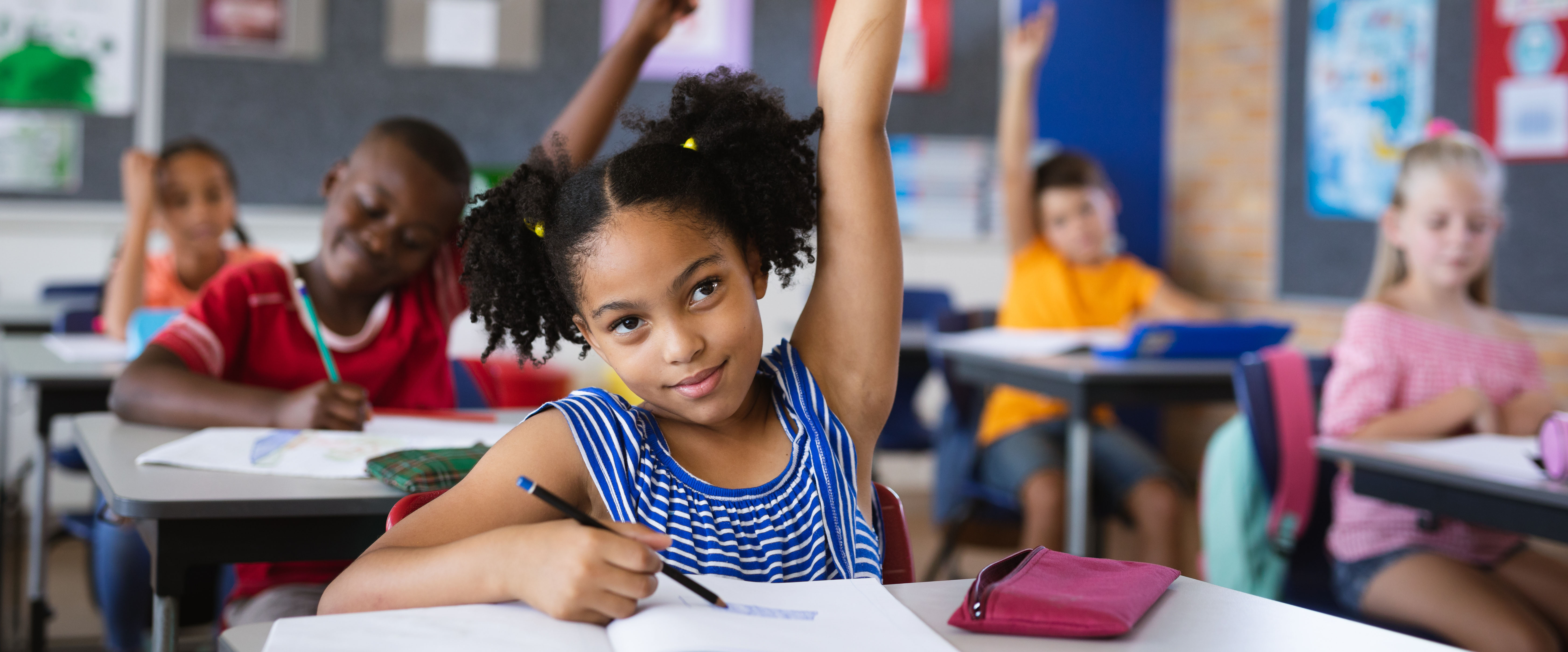 Five elementary students sitting at school desks. Three students have their hands raised.