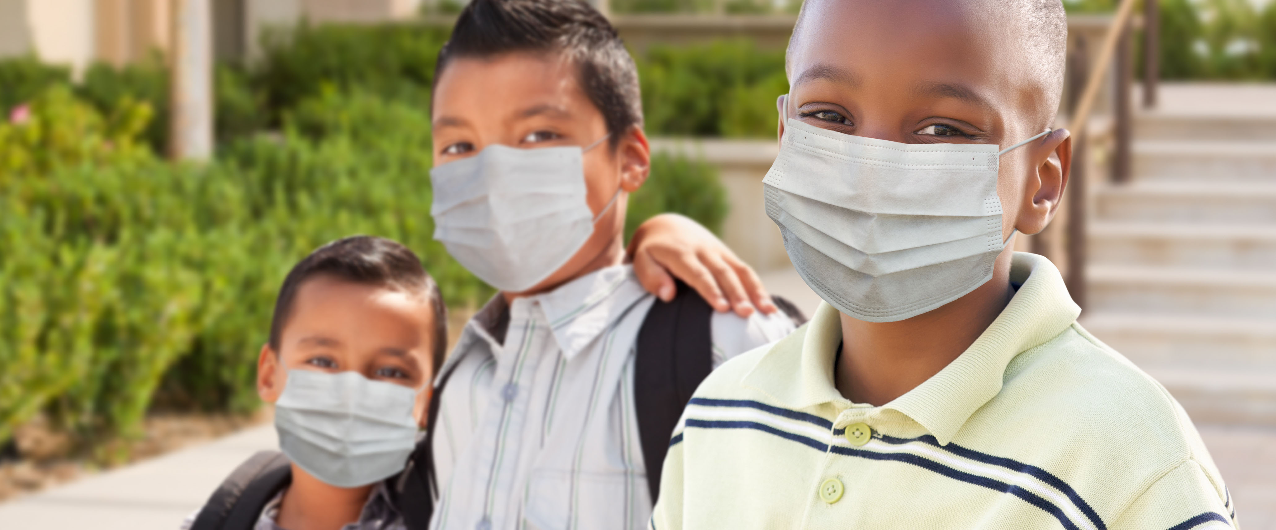 Two elementary-aged boys wearing bookbags and facemasks with their arms around each other's shoulders, standing next to another elementary-aged boy smiling wearing a face mask outside a school