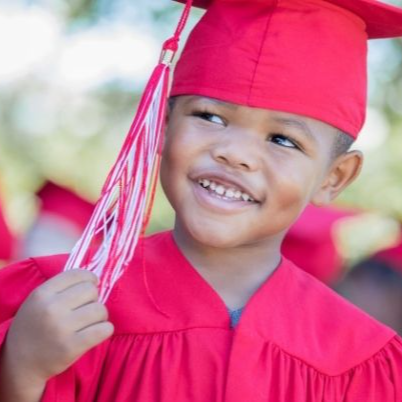 Young black boy smiling, wearing a red graduation cap and gown and touching the red and white tassel hanging from the cap.
