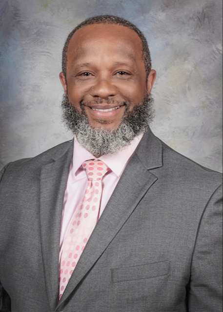Picture of Principal Cyrus Waters smiling. Black man with a black and gray beard, wearing a gray suite with a pink shirt and pink tie.