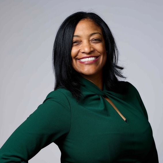 A smiling woman with black hair stands with arms at sides. She wears a dark green blouse.
