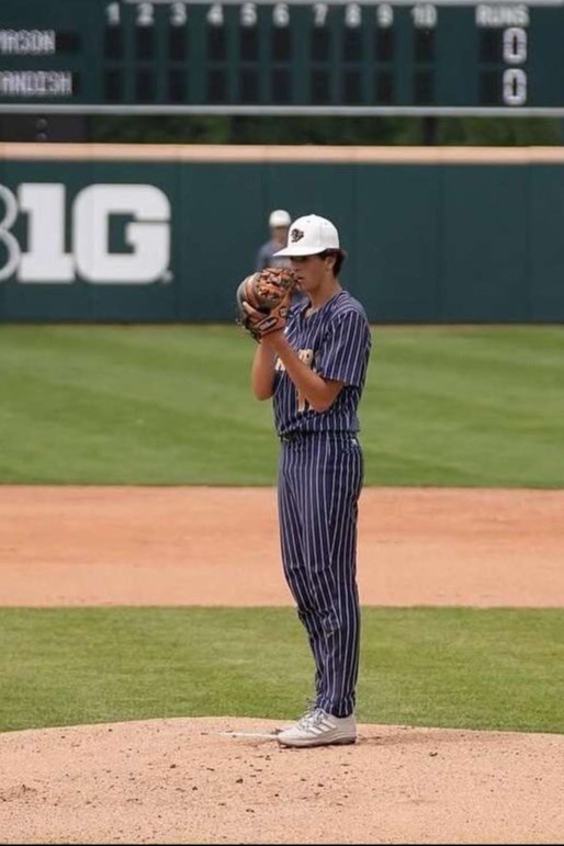 A baseball player in uniform on a mound. A scoreboard behind the player shows zeros in all columns.