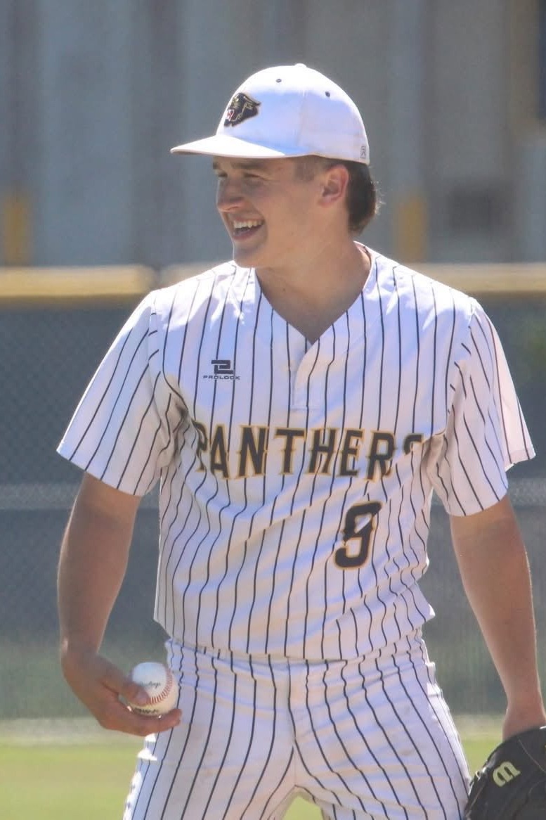 A baseball player in a "Panthers" jersey, cap, and glove, holding a ball and looking sideways on a baseball field.