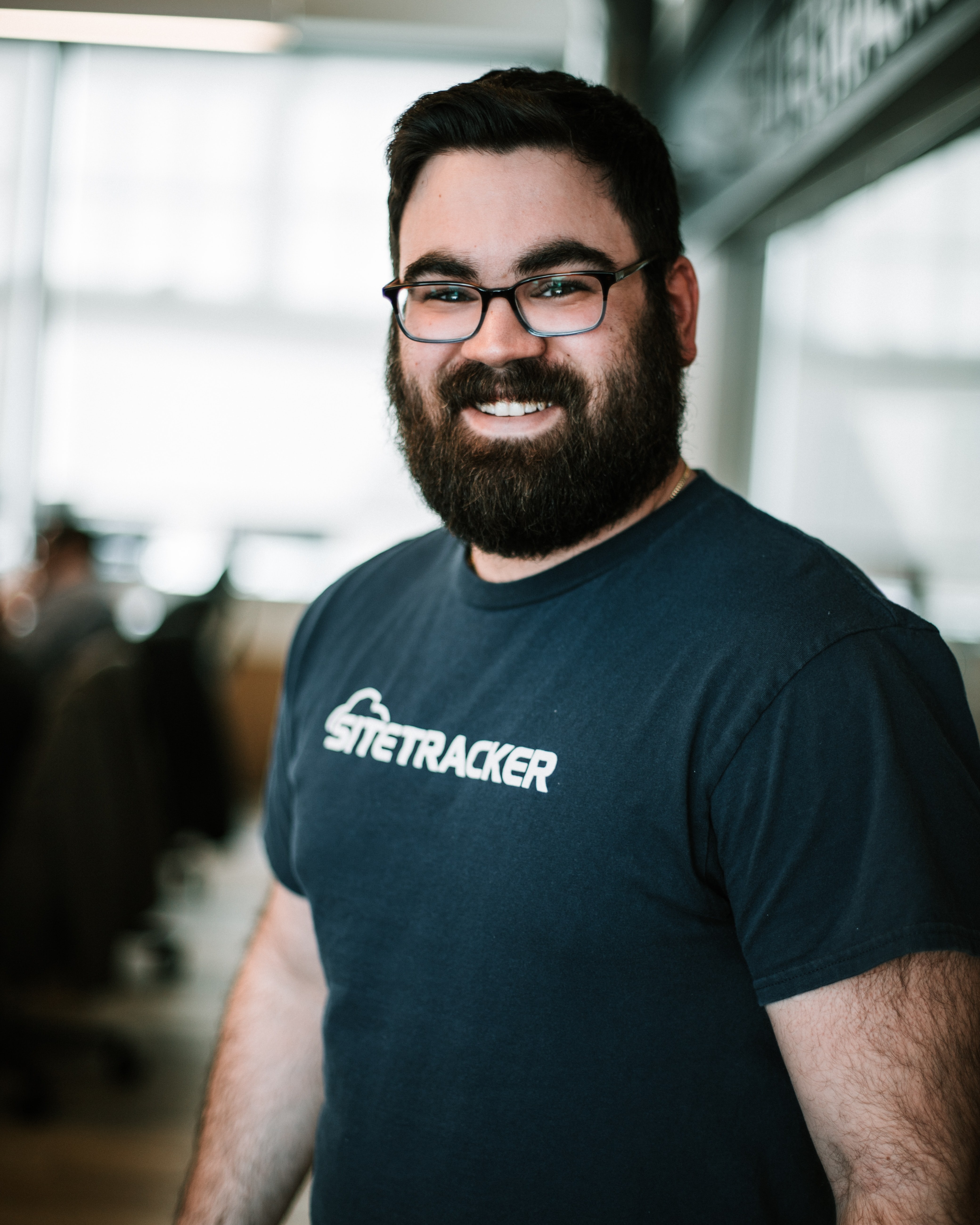 Man in glasses and beard, wearing a dark blue t-shirt with white writing, standing in an office.