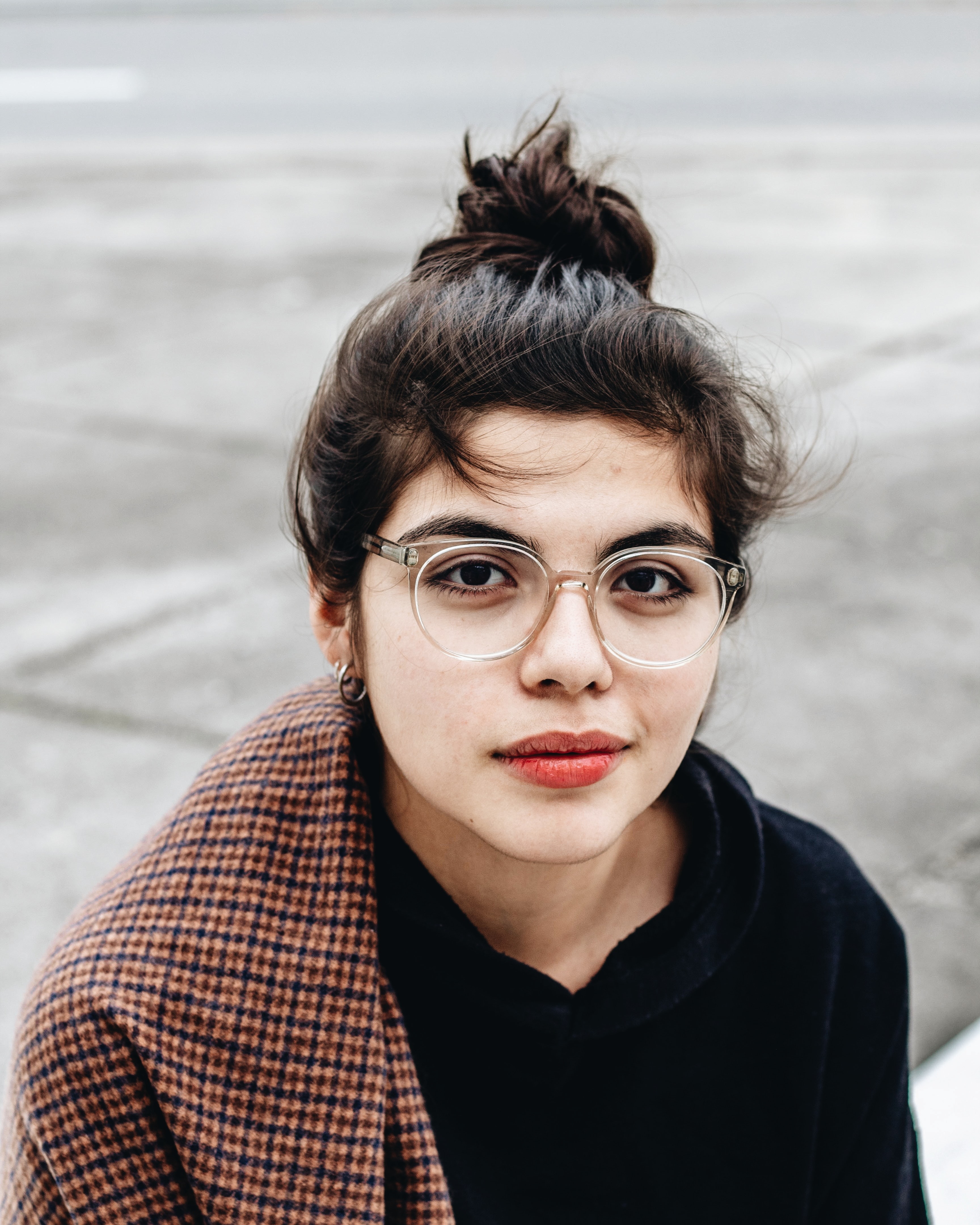 Woman with glasses, bun hairstyle, and black top. She wears a plaid coat. Behind her is a paved road.