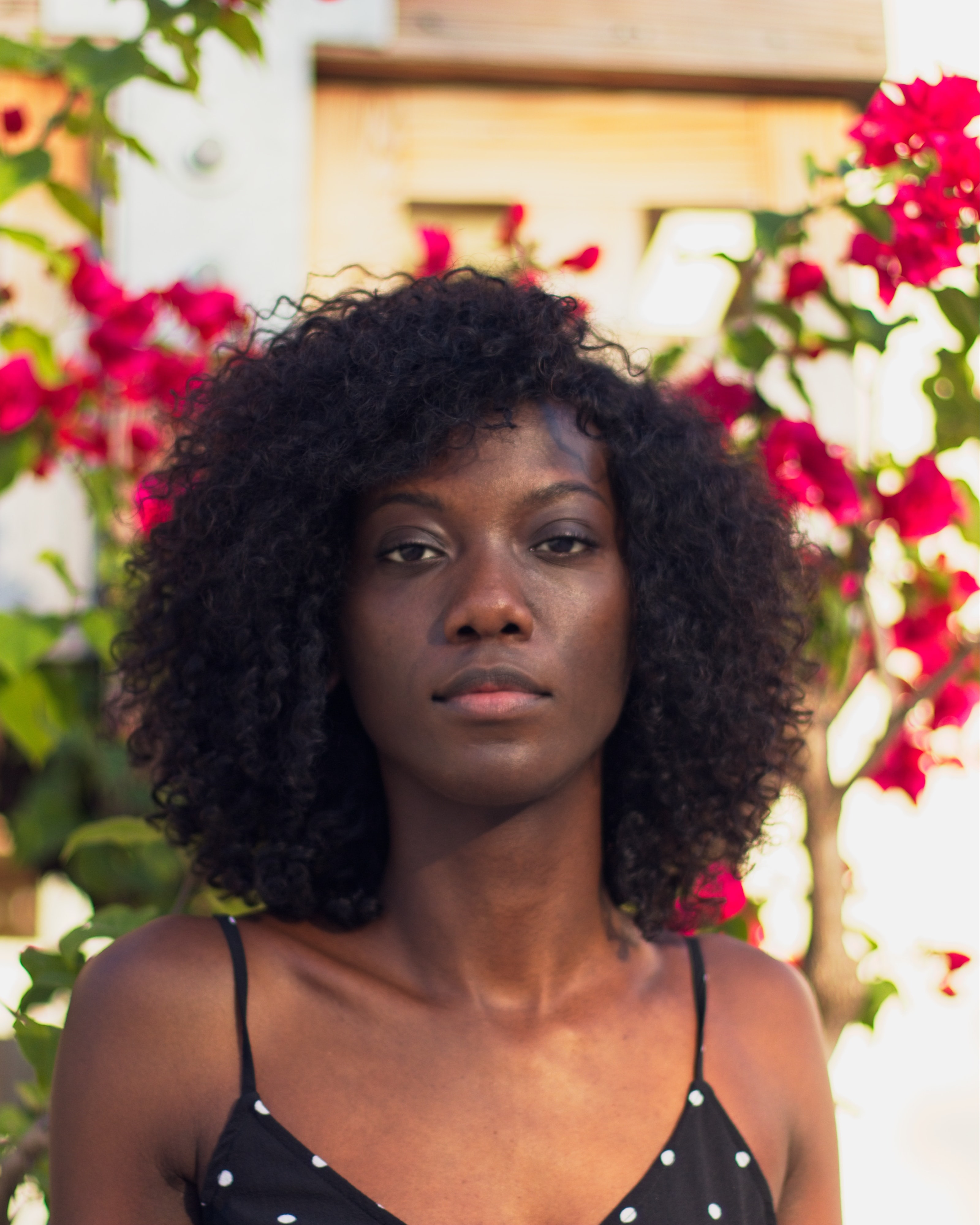 Woman with curly hair wearing a polka-dot top standing in front of a bush with red flowers.