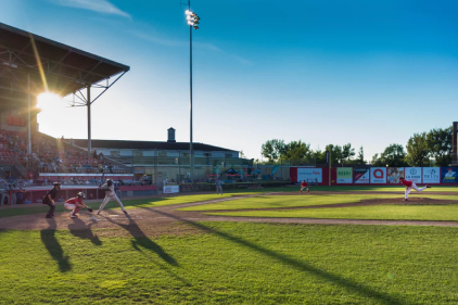 Baseball game in progress with players on field under bright sun. Stadium background with trees and building.