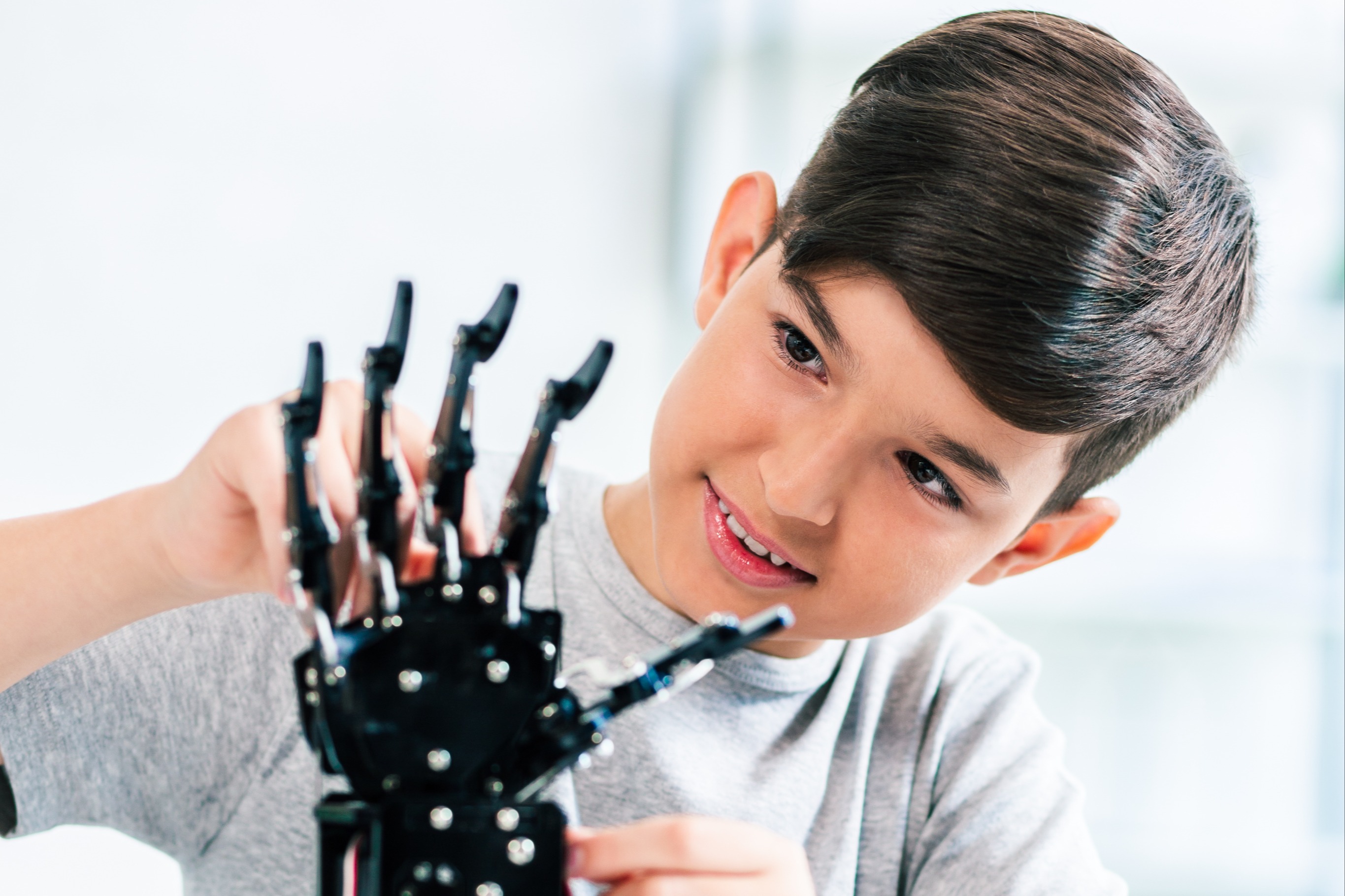 A boy wearing a gray t-shirt works on a black robotic hand with wires on a table.