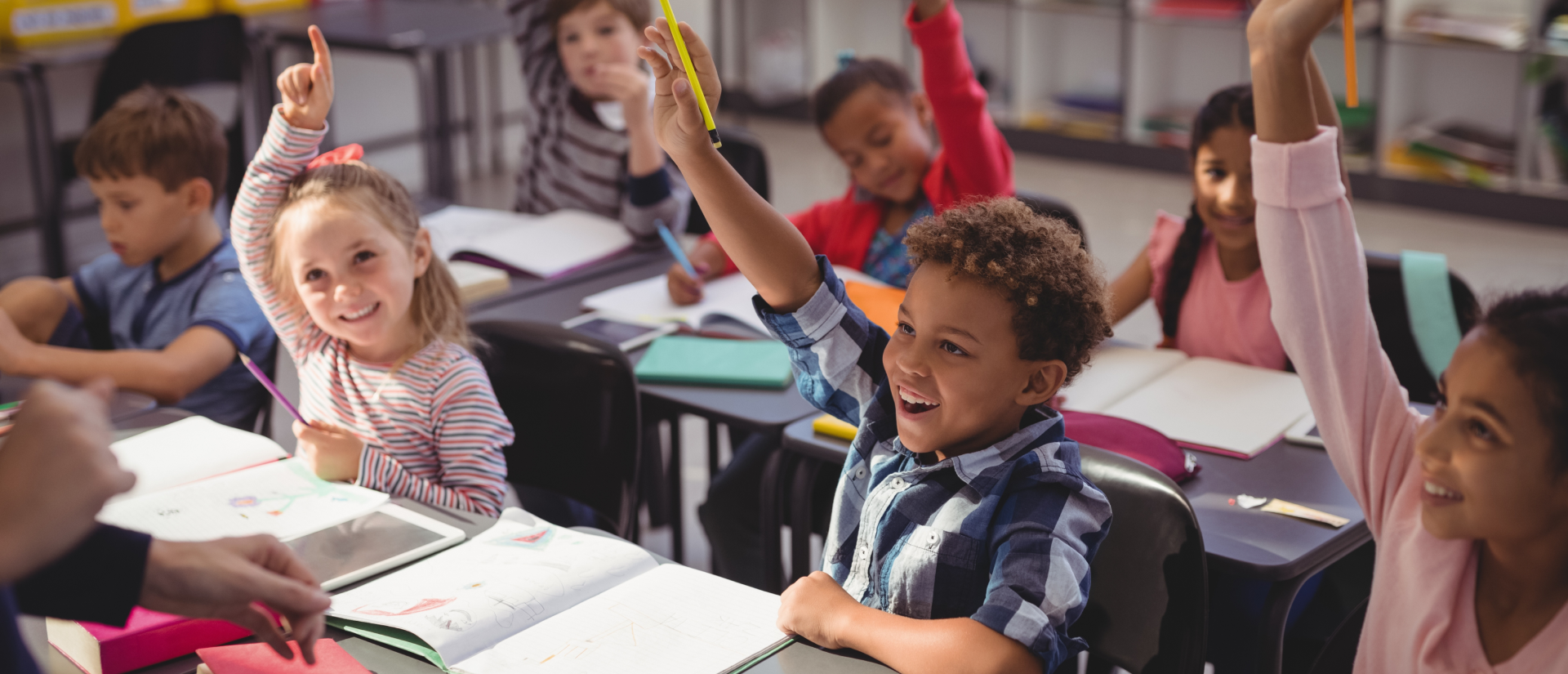 Students in a classroom raising their hands, seated at desks with various school materials.
