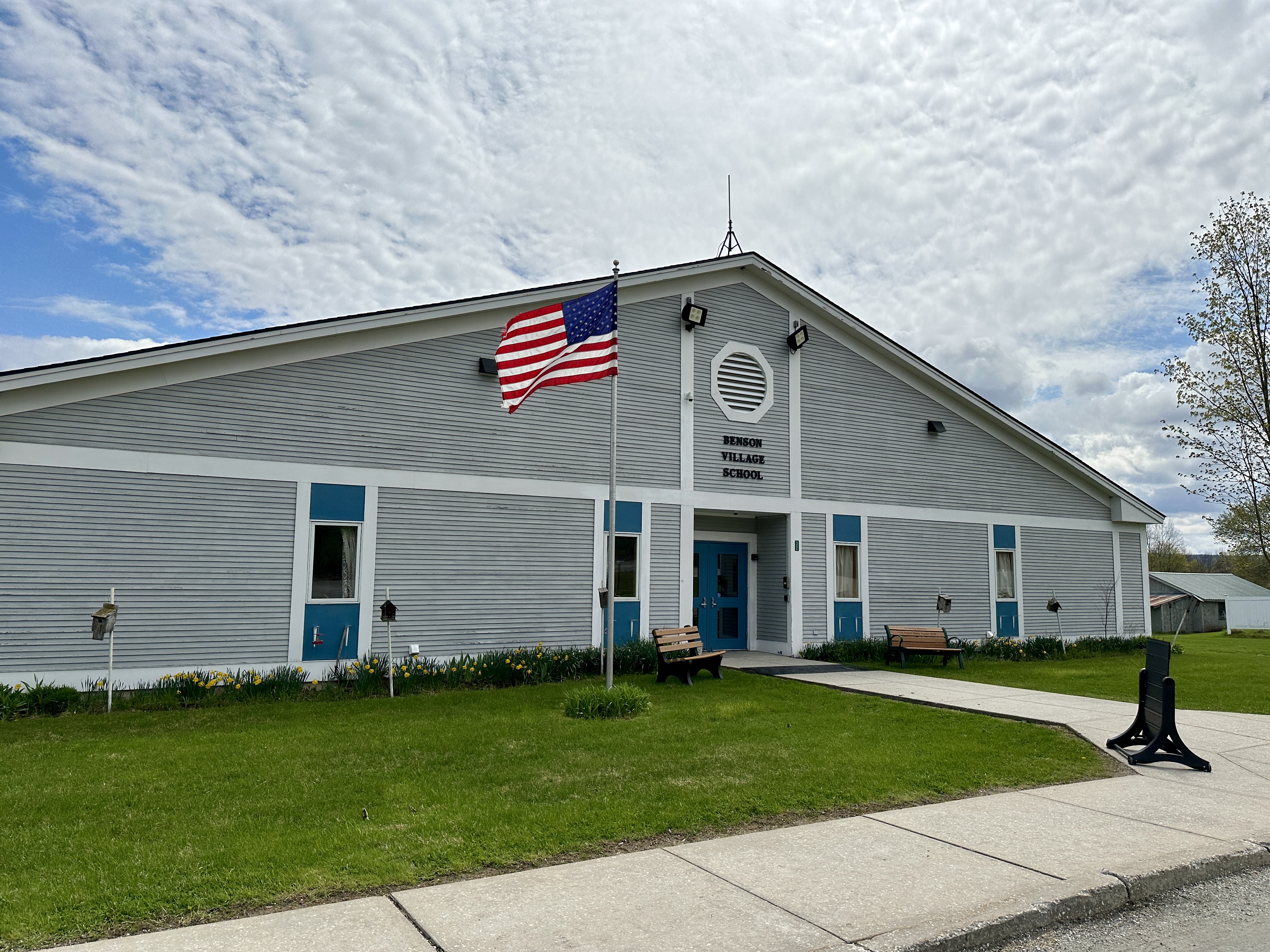 Front view of Benson Village School on a partly cloudy day, with an American flag waving on a flagpole in front of the gray and blue building.