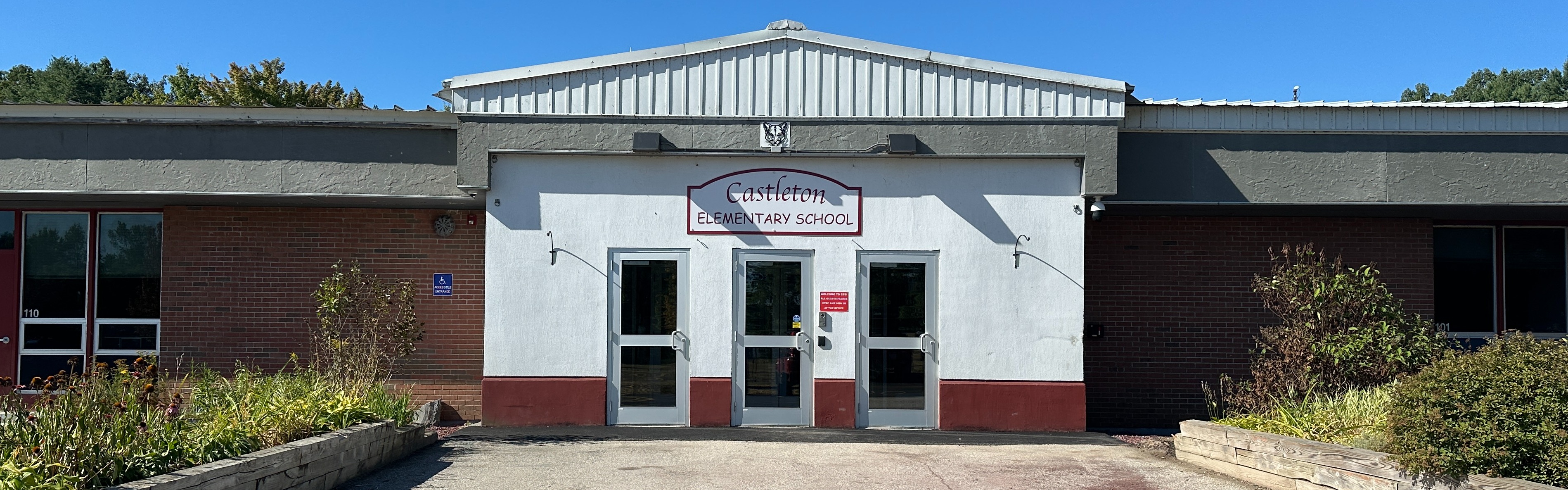 Front entrance of Castleton Elementary School, showing the school sign above 3 doors. Some clouds in a blue sky.