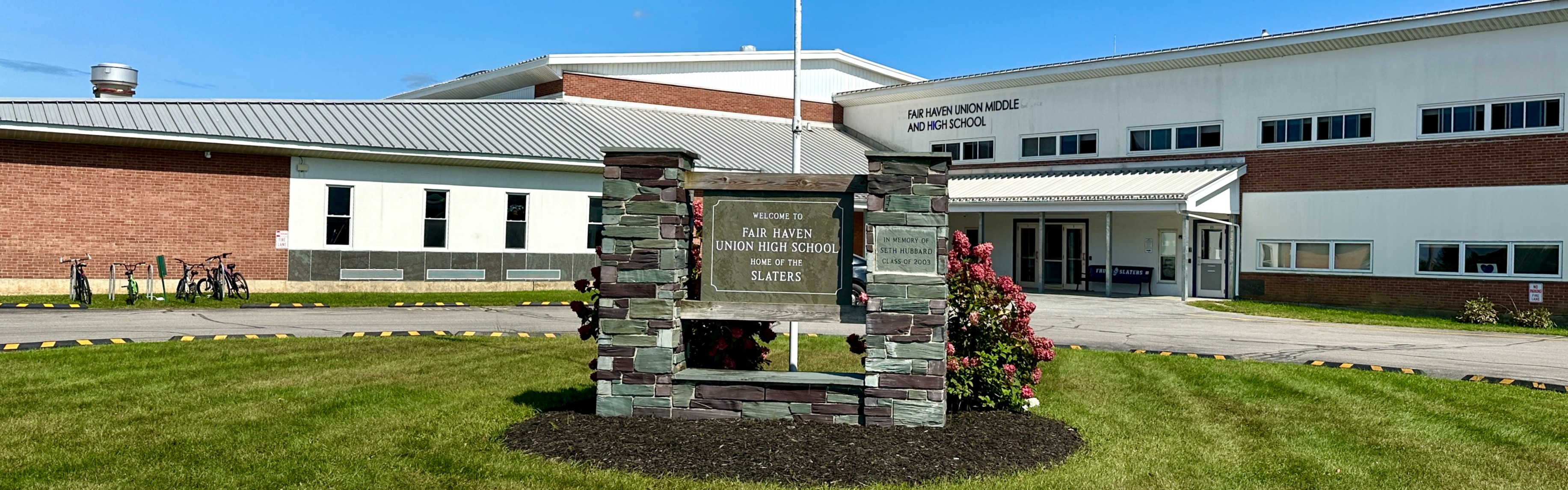 Front of the Fair Haven Union Middle and High School, with a flag post and slate sign in front of it, and a sunny, blue sky