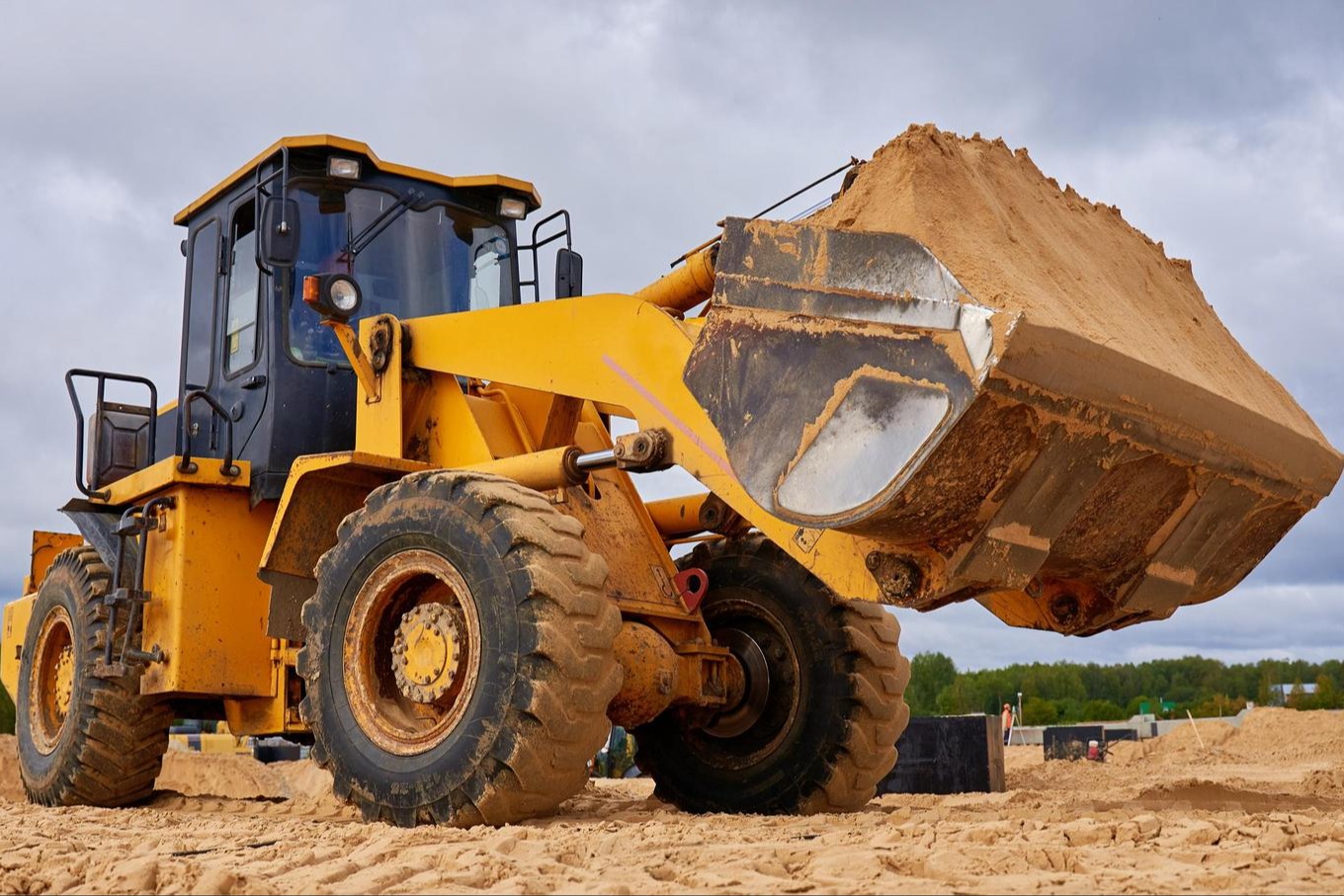 A yellow bulldozer with a large bucket of dirt, on a dirt field under a cloudy sky.