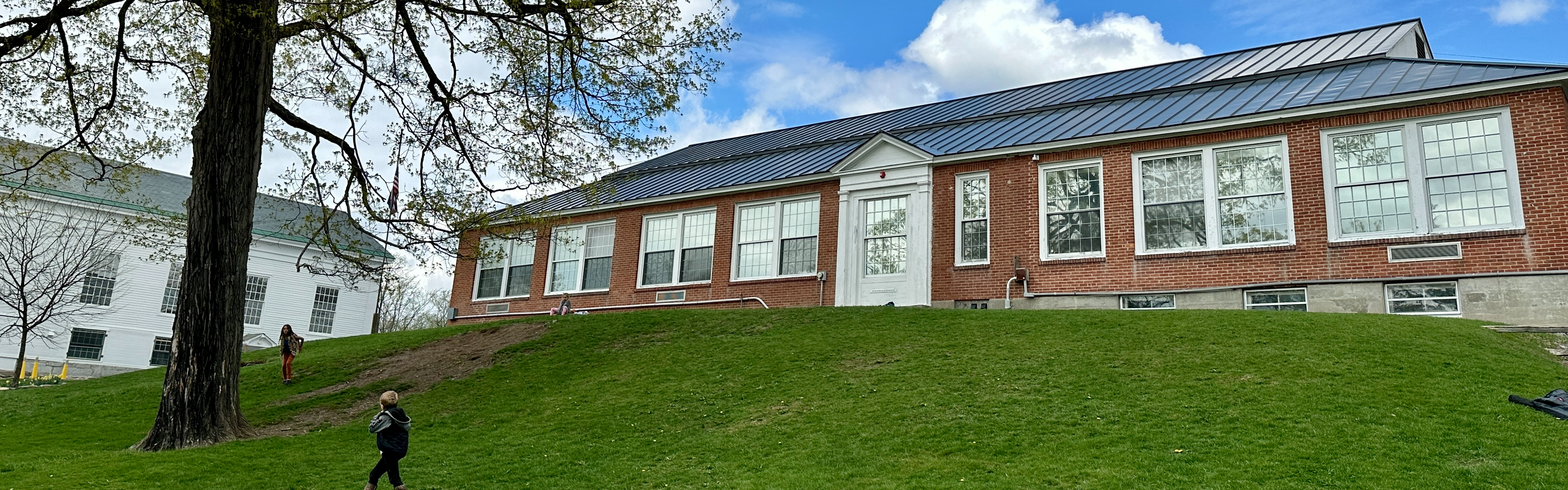 Photo of Orwell Village school with a blue sky and two students running down the hill in front of it.
