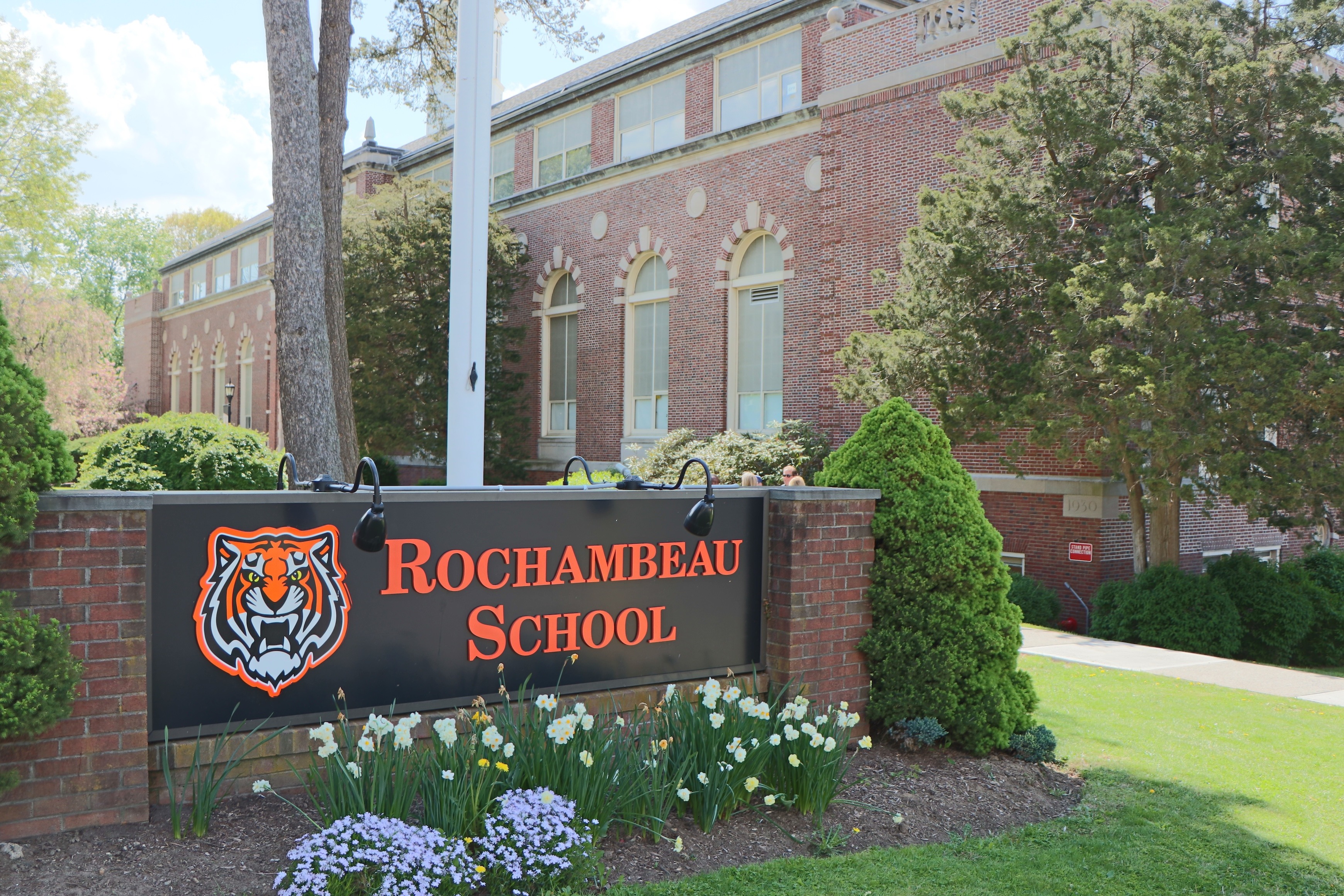 Sign for Rochambeau School with a tiger emblem. Brick building, arched windows, greenery, and flowers.