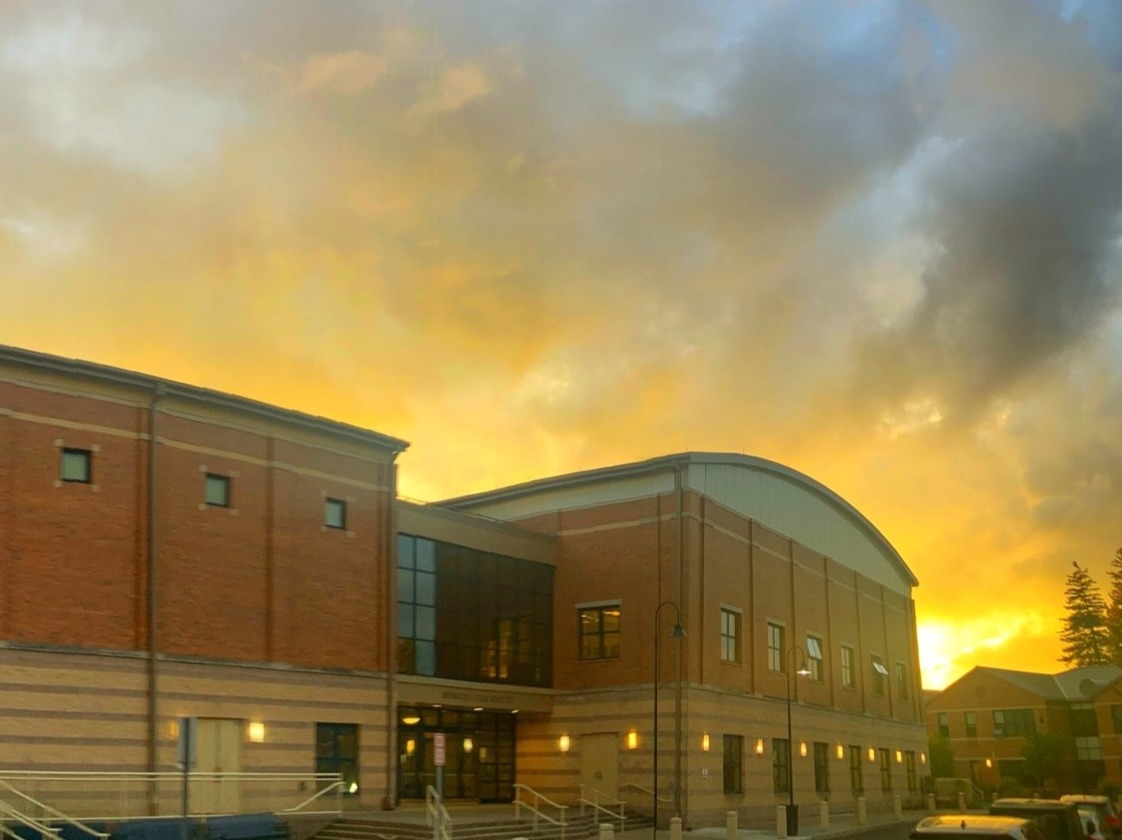 A building with brick walls and glass windows stands against a sunset sky. The sky is filled with clouds.