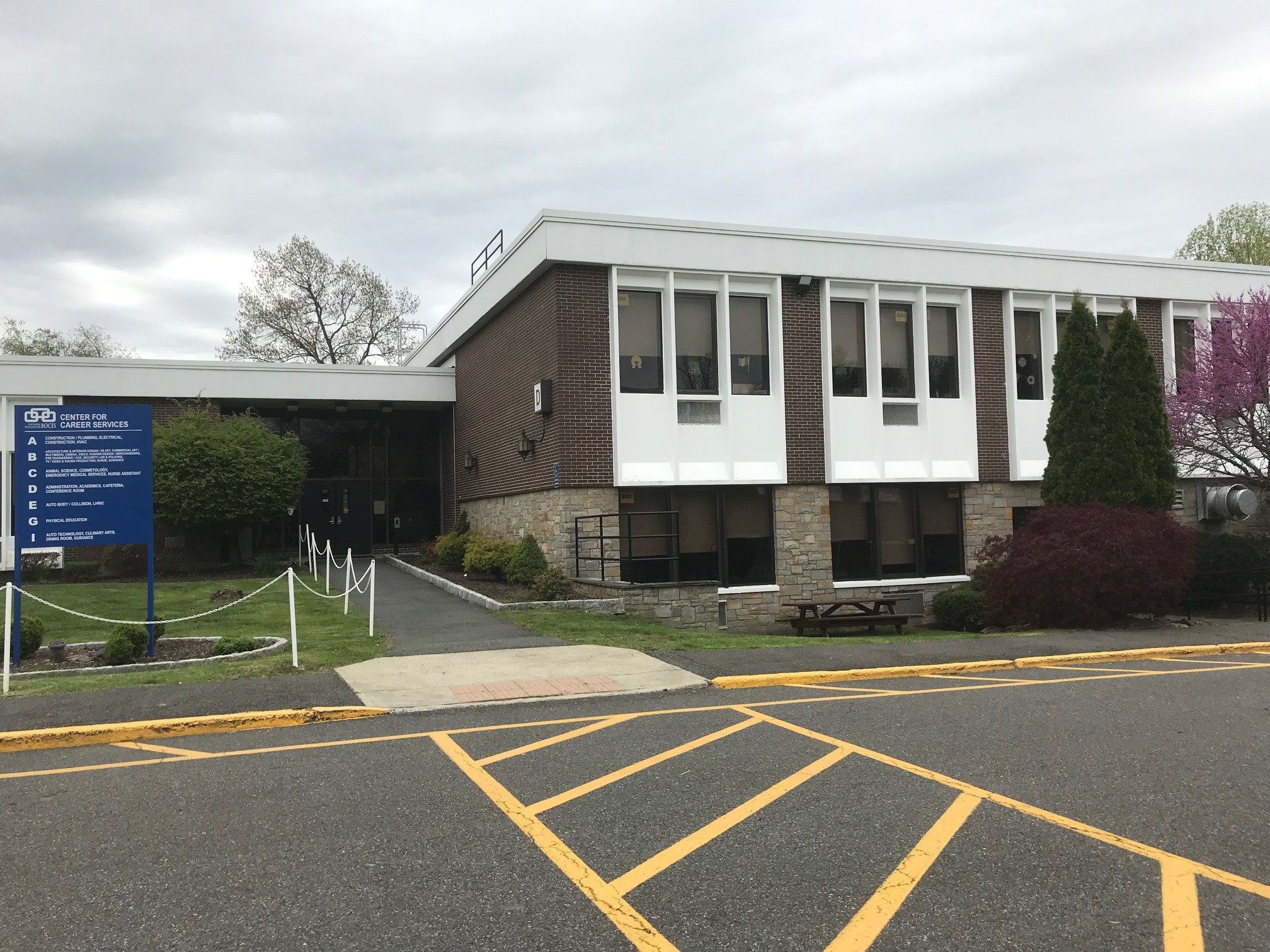 A building with many windows, a brown and white exterior, a blue sign, a parking lot, and trees.