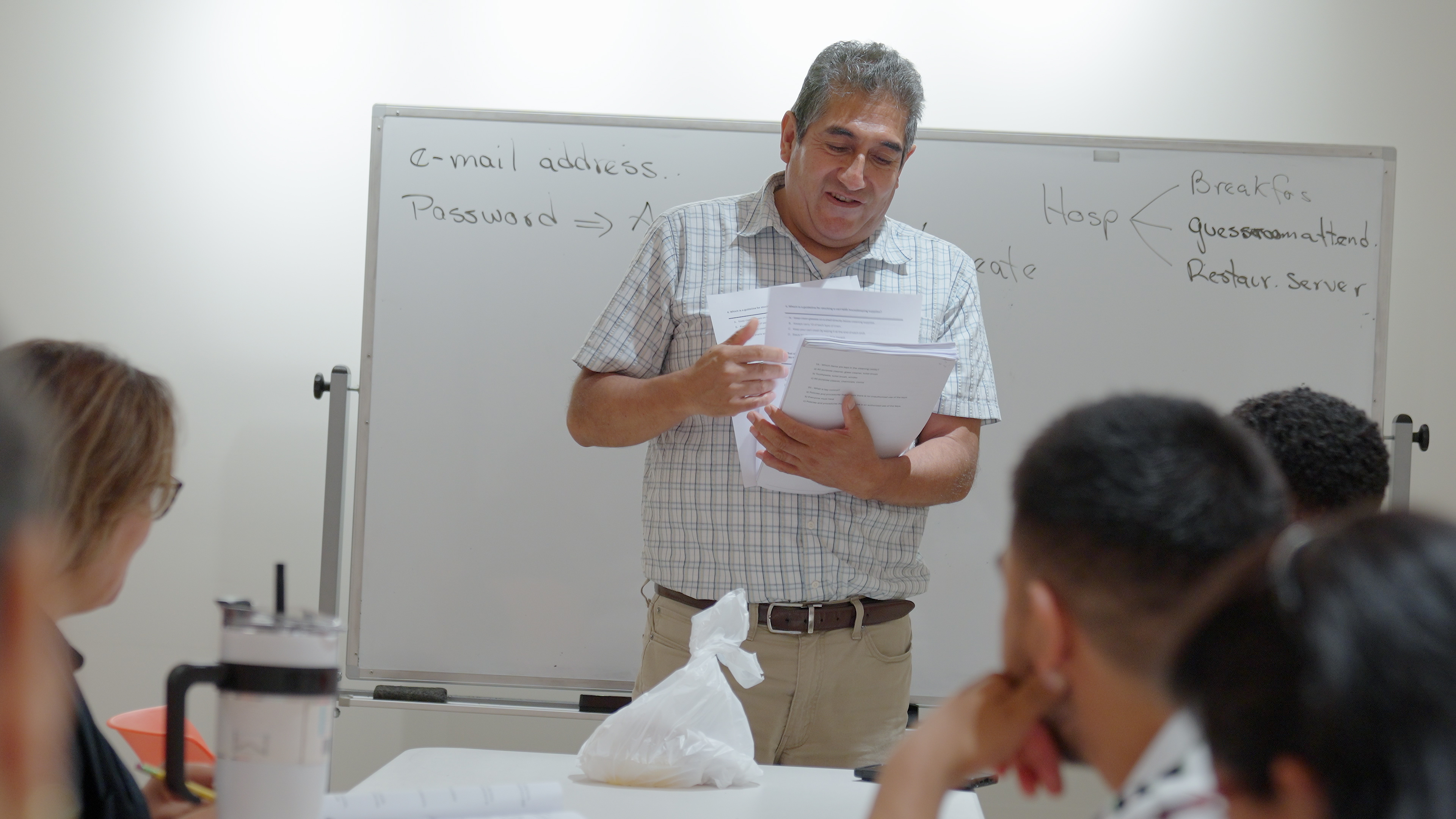 A man in a checkered shirt stands before a whiteboard, speaking to a group of seated individuals.