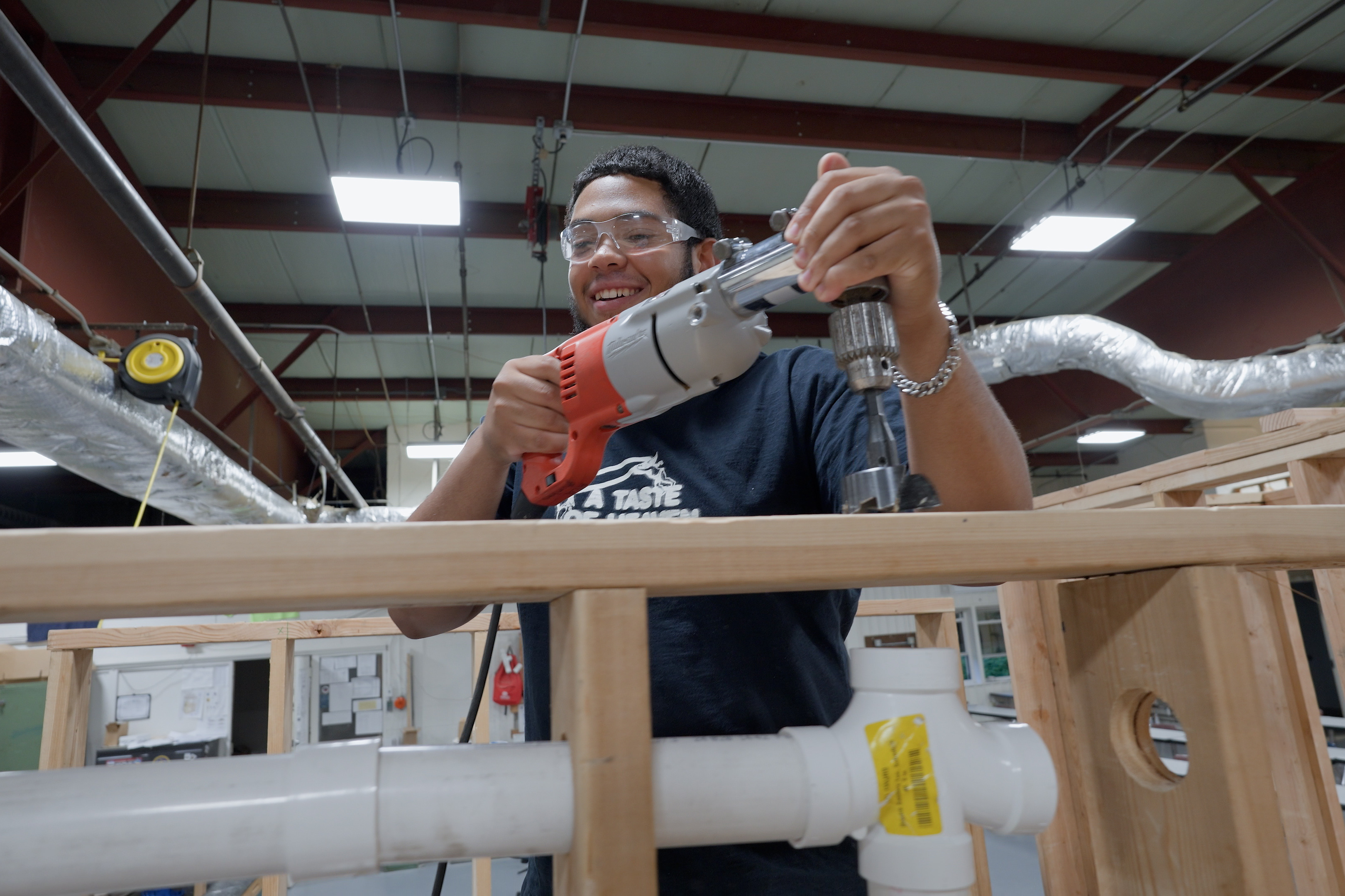 A man uses a drill while wearing safety goggles and a shirt in an industrial setting.