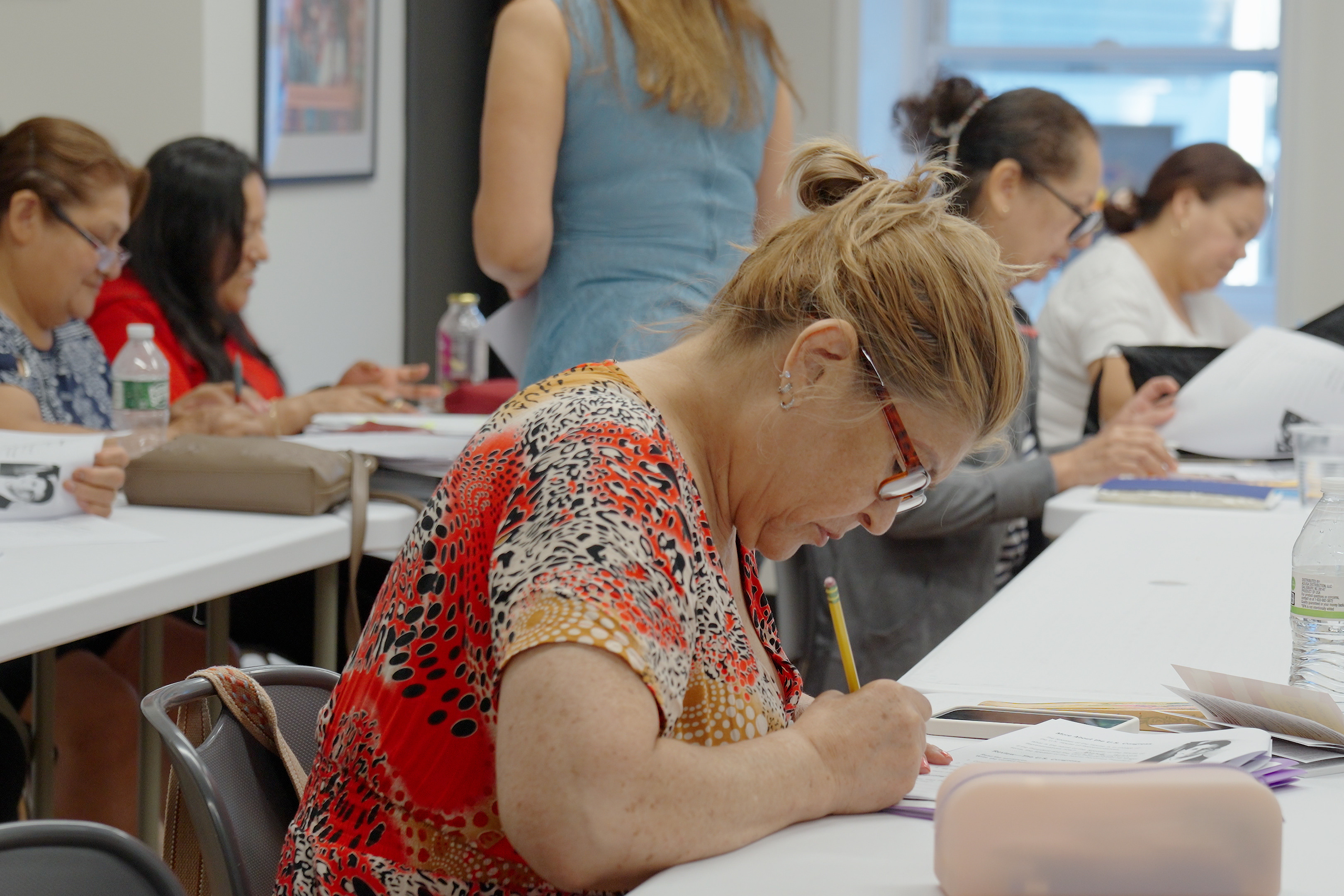 A group of people in a classroom are seated at tables. They are looking down and writing.
