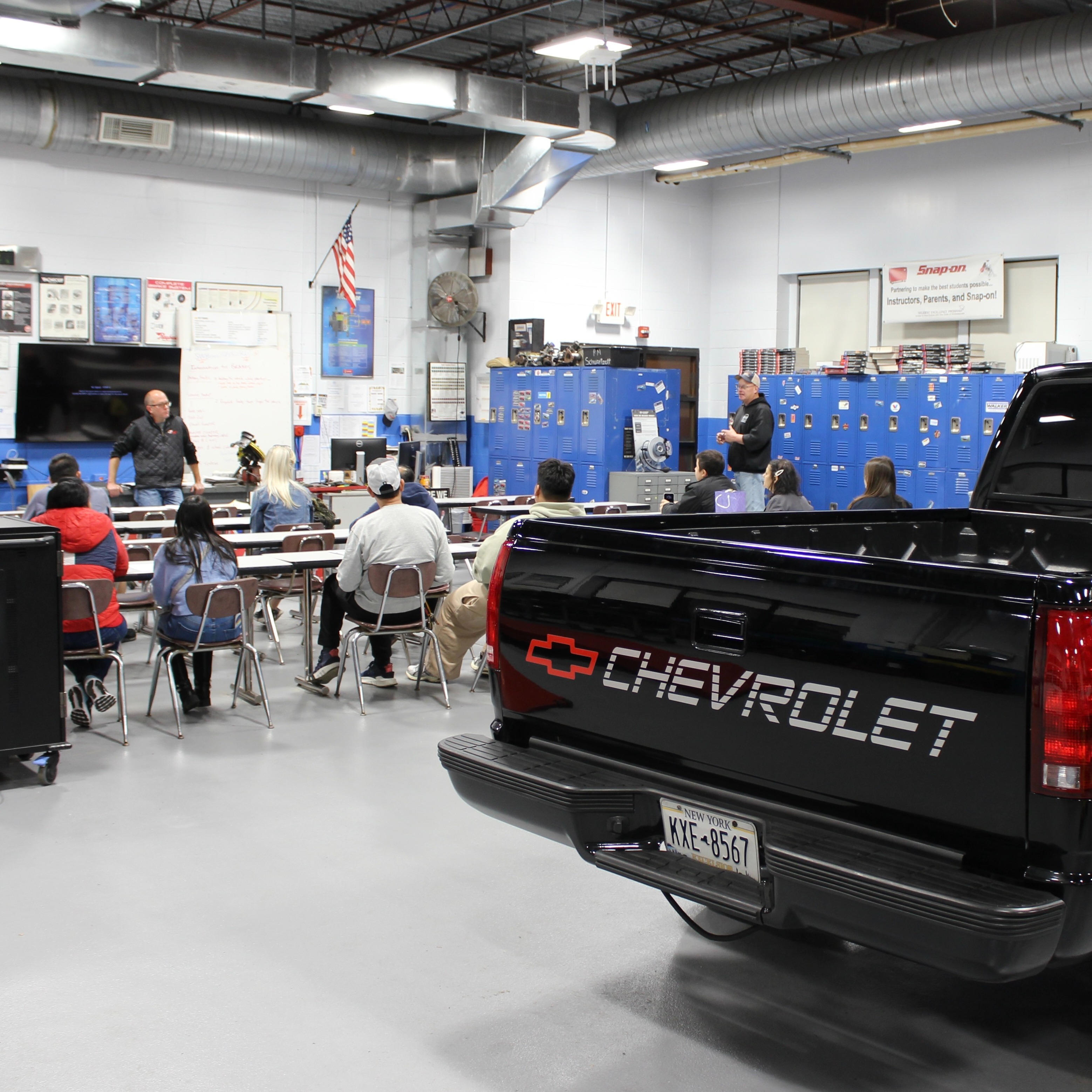 A Chevy pickup truck is parked in an auto tech classroom as the teacher lectures students in the background