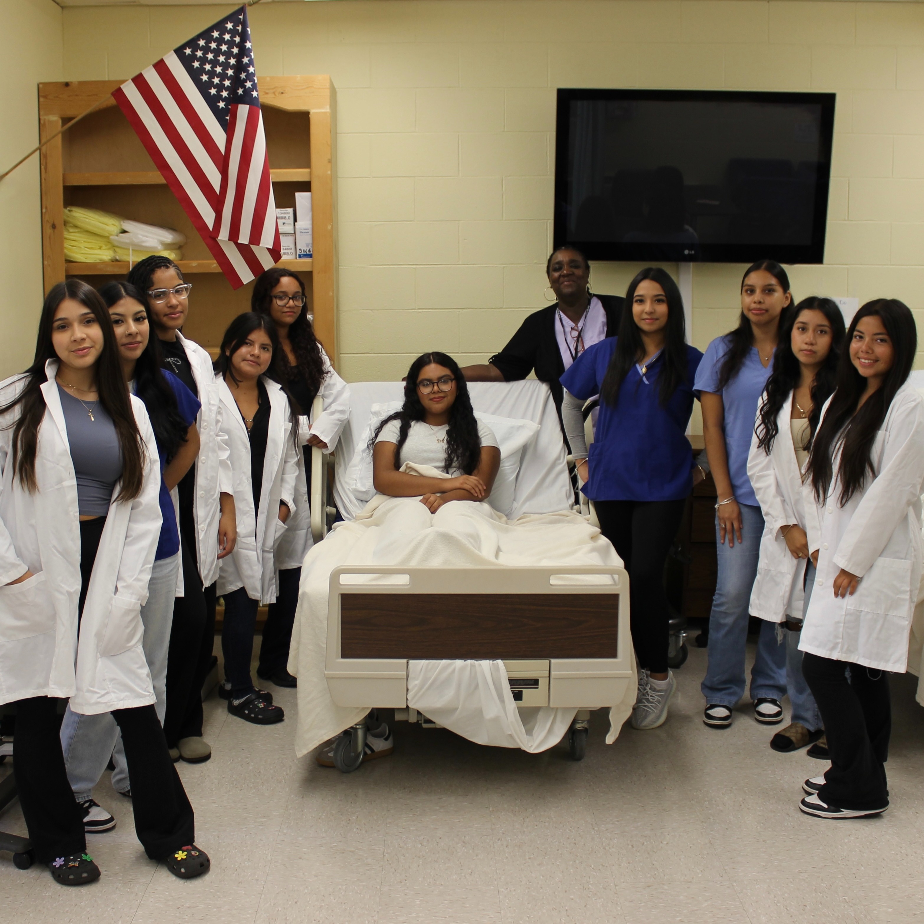 Students in white lab coats stand around a hospital bed, a man is standing behind it.