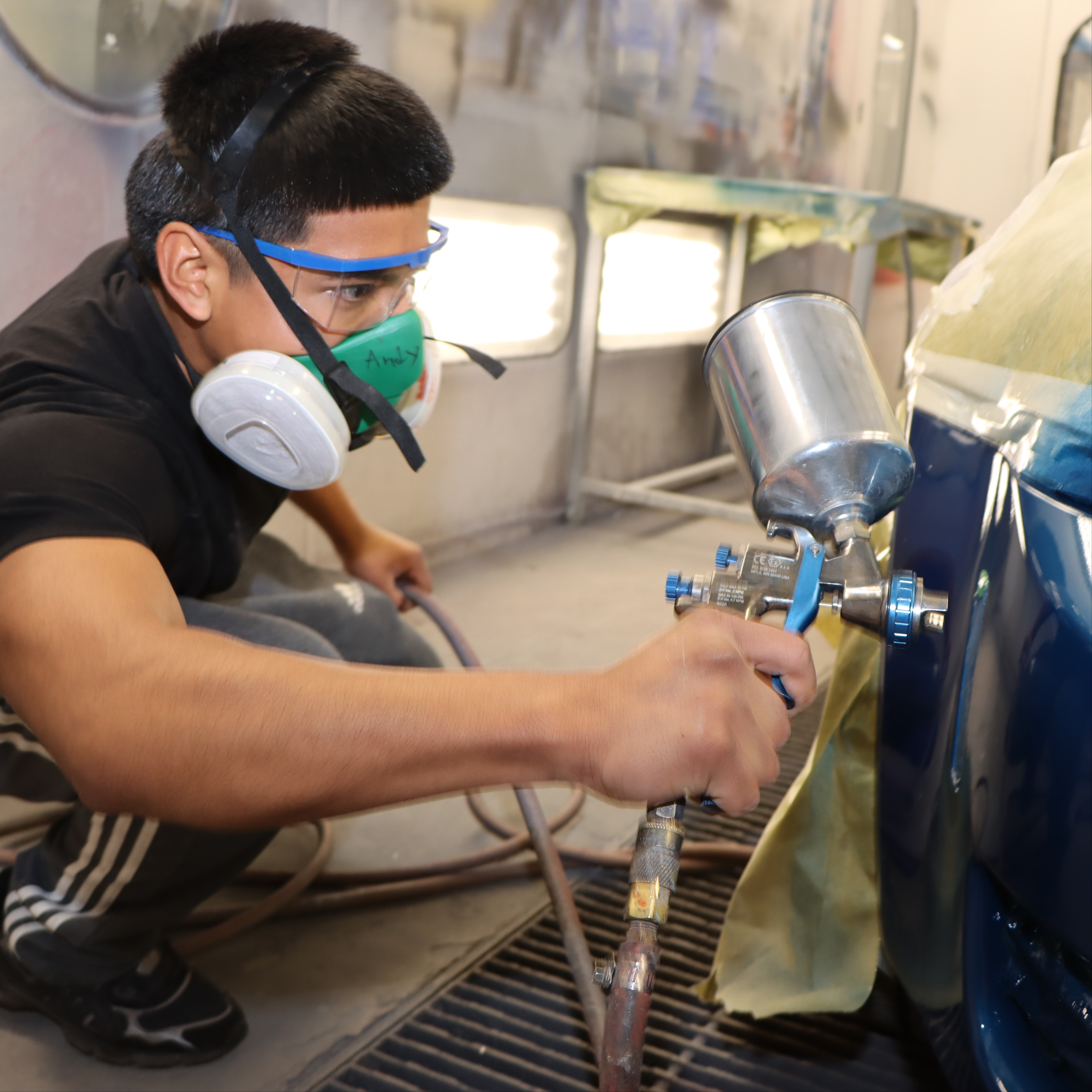 auto body student applies paint to a car using a pneumatic sprayer while wearing protective gear