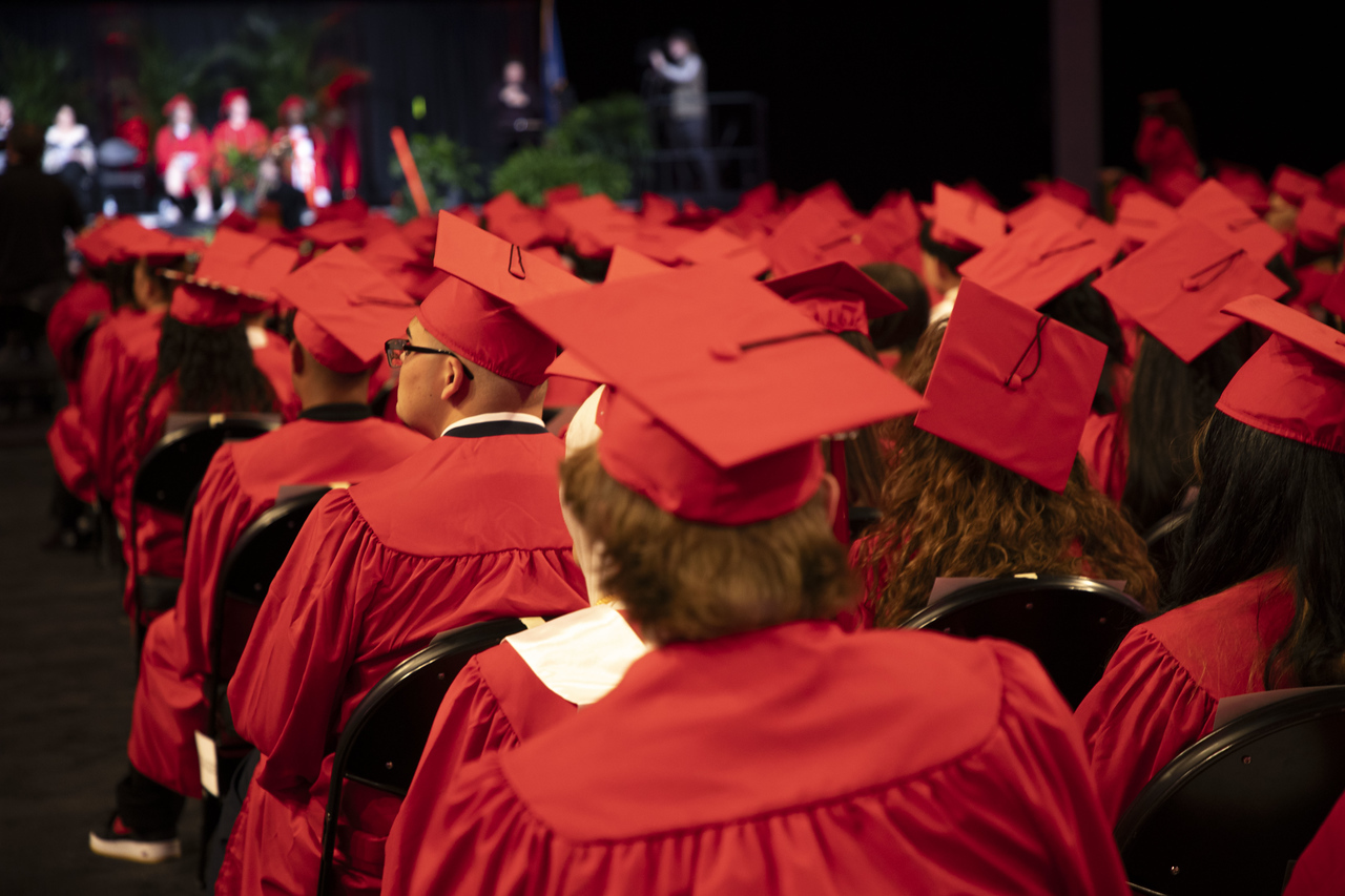 Dozens of graduates are seated in rows, viewed from behind, wearing vibrant red caps and gowns. Their backs face forward, attending a ceremony with blurred figures visible on a distant stage against a dark background.