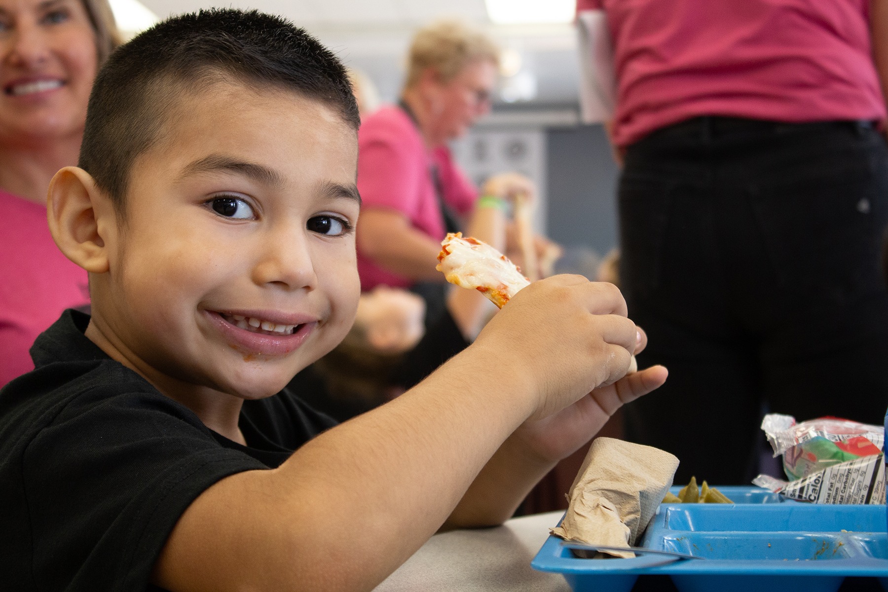 Elementary student eating pizza and smiling at the camera.