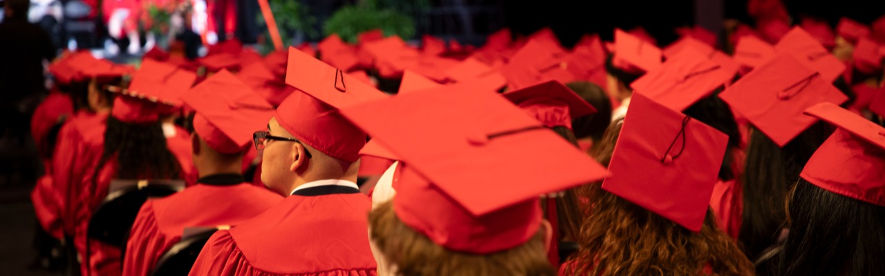 Dozens of graduates are seated in rows, viewed from behind, wearing vibrant red caps and gowns. Their backs face forward, attending a ceremony with blurred figures visible on a distant stage against a dark background.