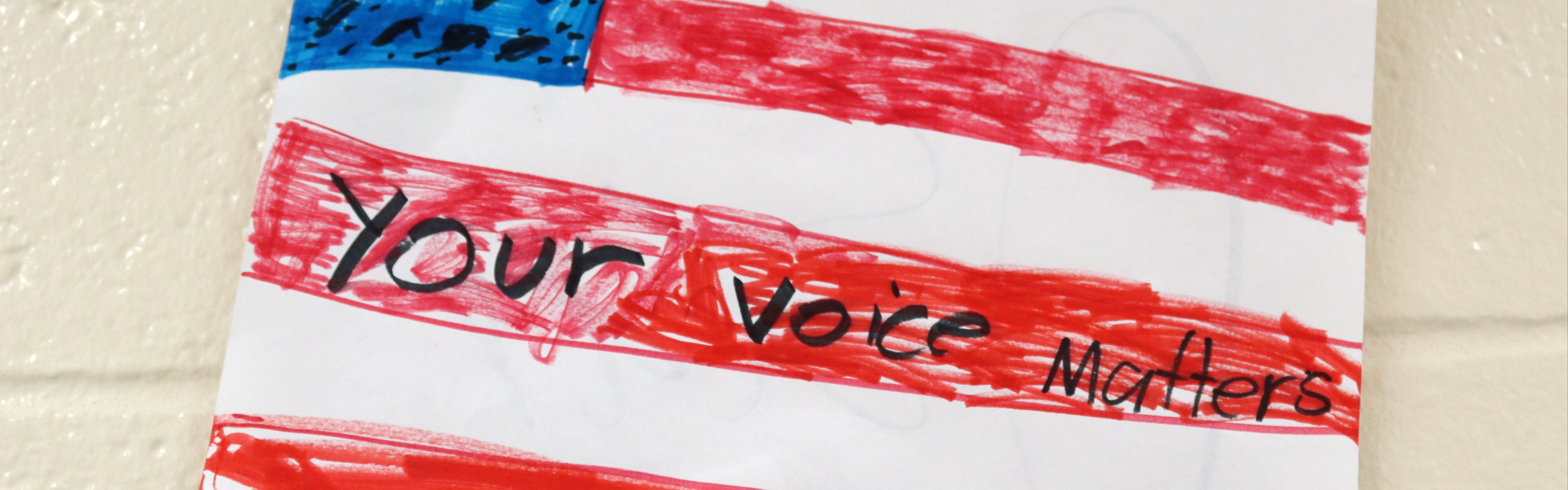 A hand-drawn American flag in red and blue marker on white paper, taped to a cream-painted cinder block wall. Handwritten black lettering across the stripes reads "Your voice matters."
