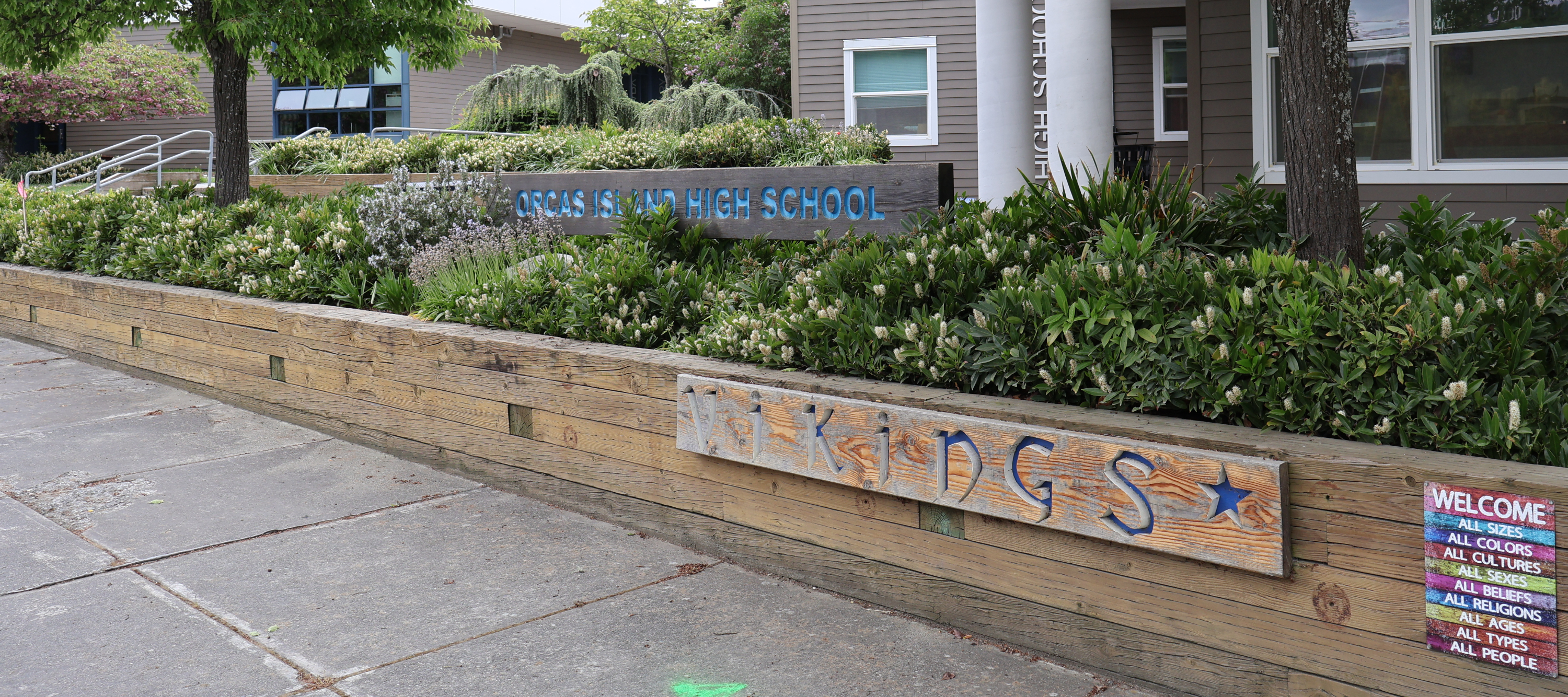 A wide outdoor shot of the entrance area of Orcas Island High School. A wooden retaining wall features a carved sign reading "VIKINGS," and a smaller rainbow-colored sign welcoming all people. Behind it, lush green landscaping surrounds a larger sign that reads "ORCAS ISLAND HIGH SCHOOL."