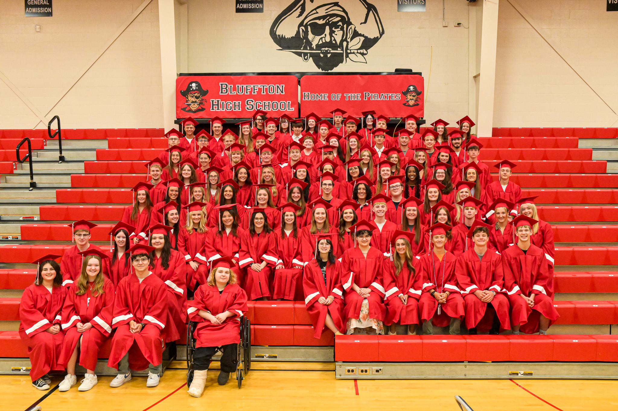 A group of graduates in red gowns and caps pose for a photo on a gymnasium bleacher.