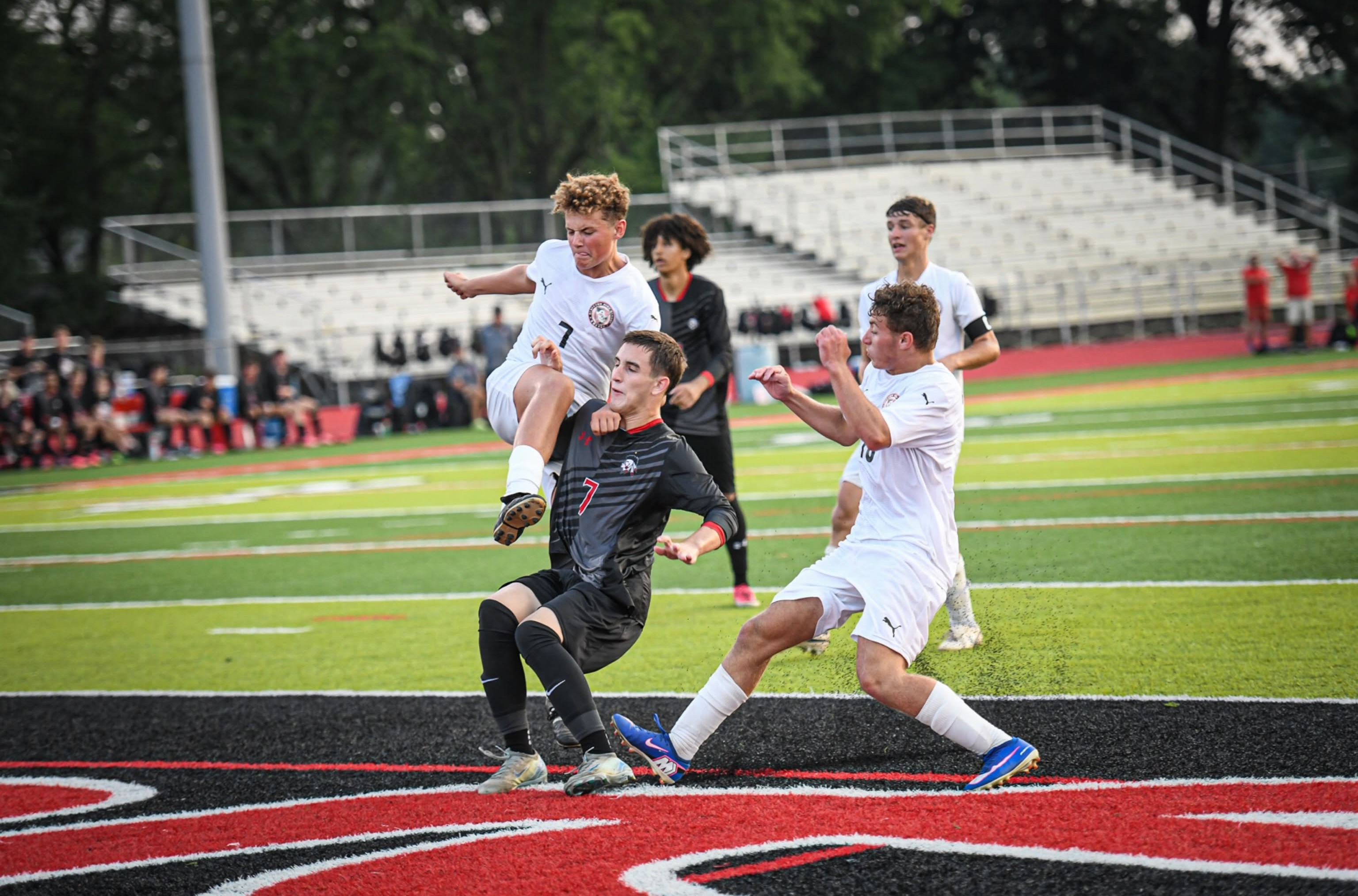 Players in white and black uniforms compete on a field with a red and black logo. One player leaps mid-air.