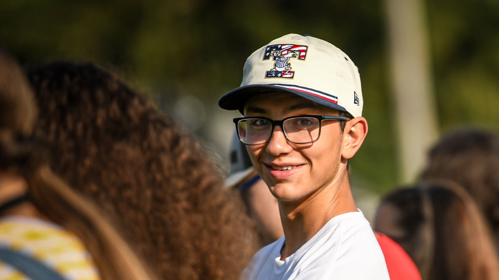 A man wearing a white hat, glasses, and a white shirt stands among others in an outdoor setting.
