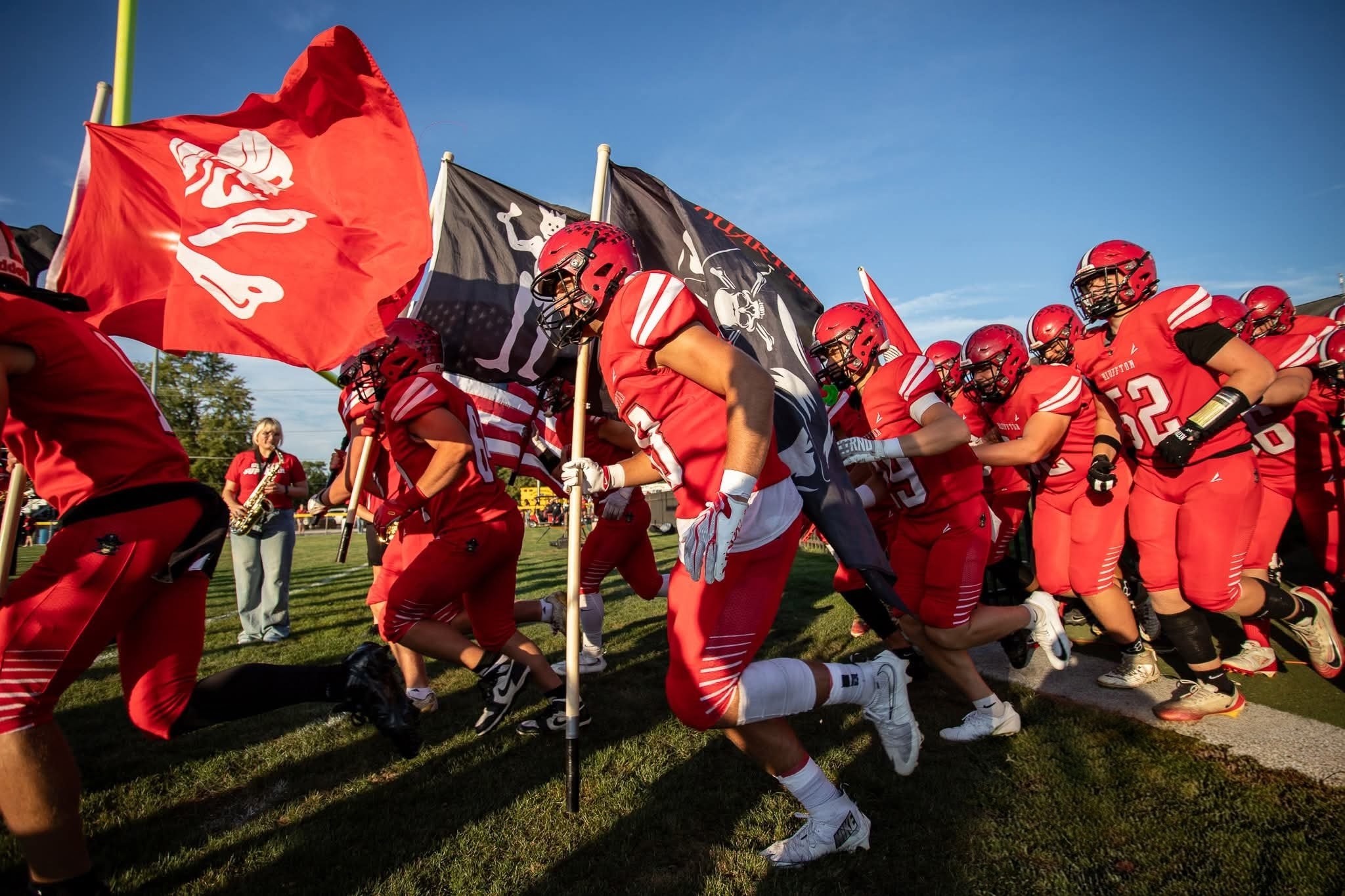 Football team in red uniforms runs onto the field carrying flags and holding poles. One person carries an American flag.
