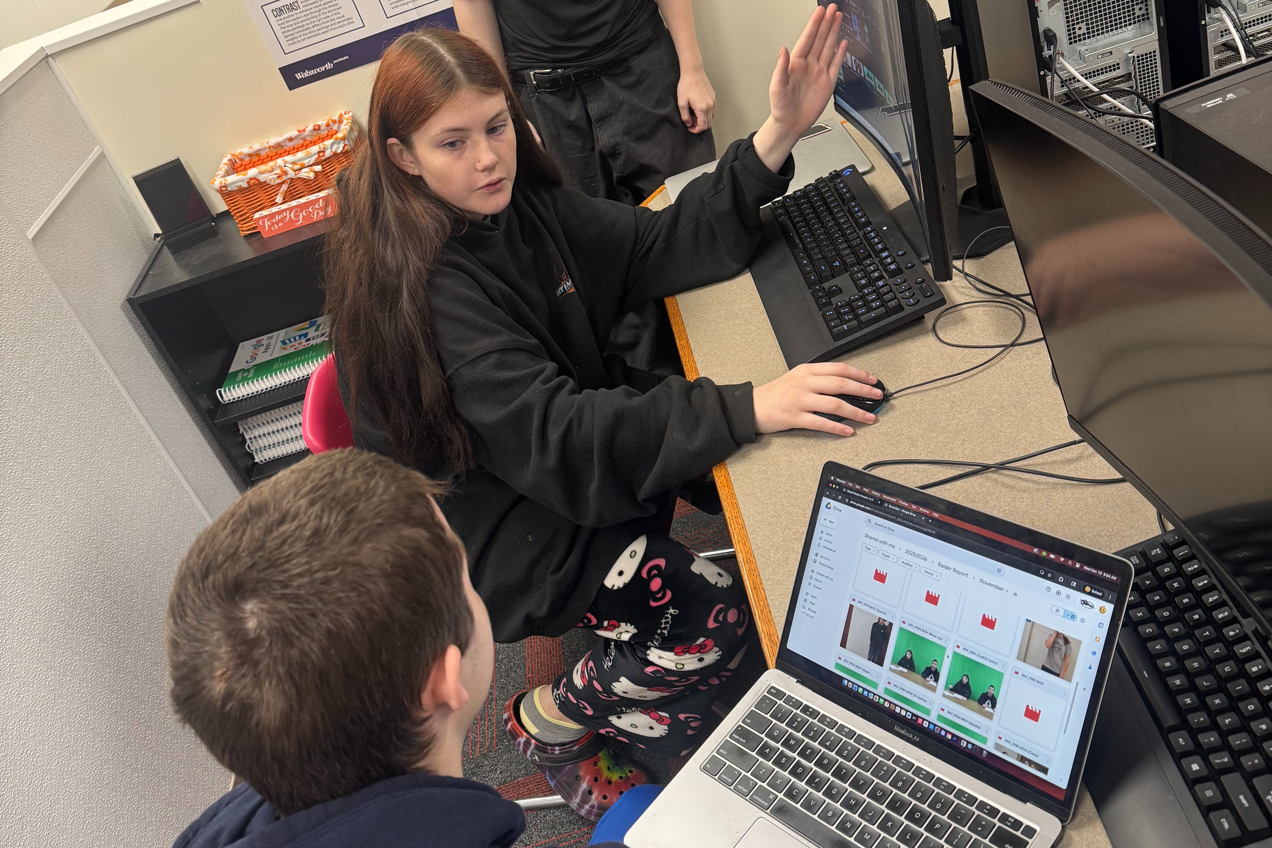 Three students work together in a computer lab. One student sits at a desktop computer, another sits beside them using a laptop displaying photo thumbnails, and a third student stands behind them observing. A “Design Rules” poster hangs on the wall, and a small sign on a computer tower reminds students to clean up and take care of their workspace.