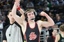 A high school wrestler in a black and red singlet celebrates a victory as the referee raises his arm. The wrestler smiles and pumps his fist while the crowd watches in the background.