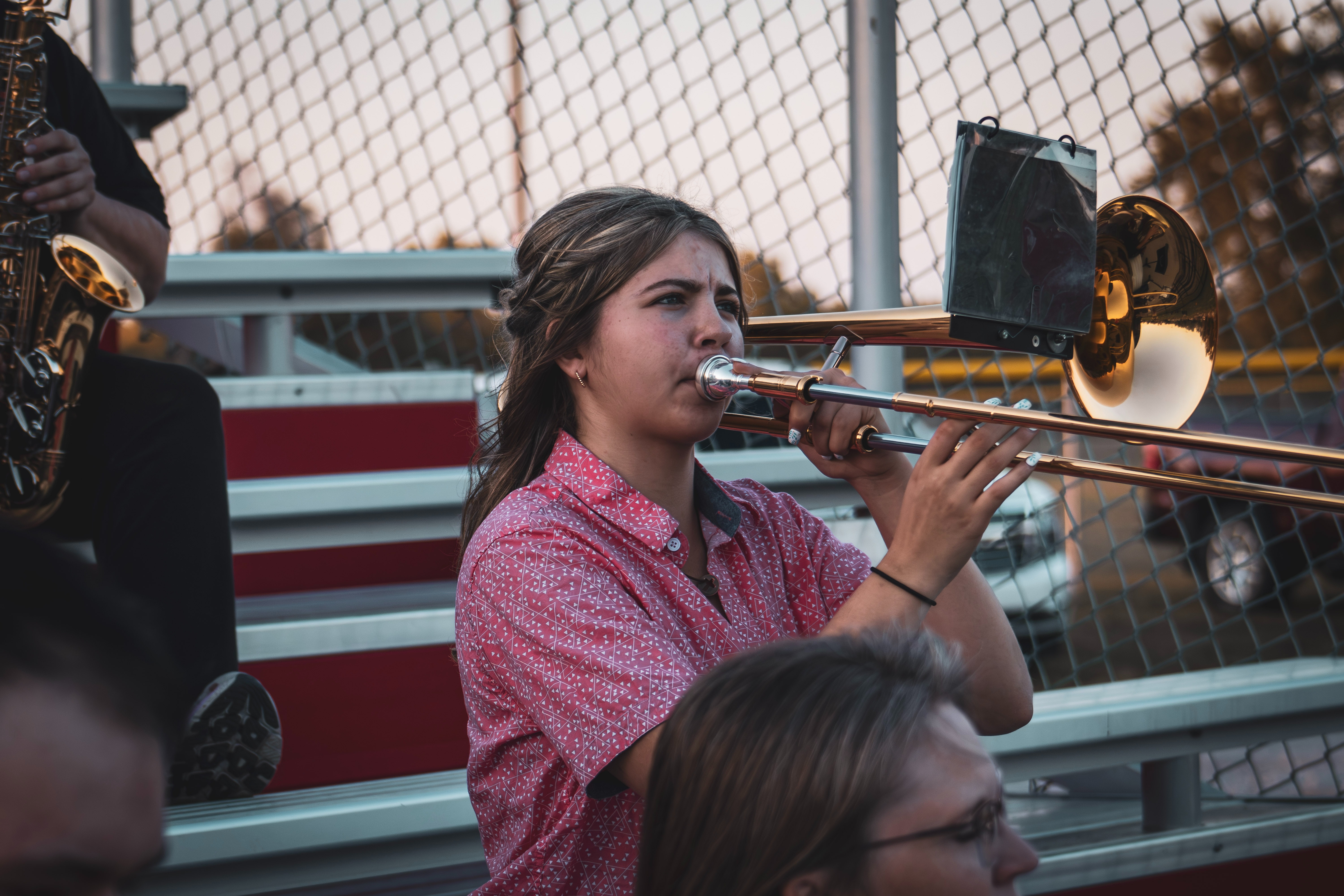 A student plays the trombone in the bleachers during an outdoor event. She wears a patterned red shirt, and a music lyre is attached to the instrument. Another band member is partially visible nearby, and a chain-link fence lines the background.