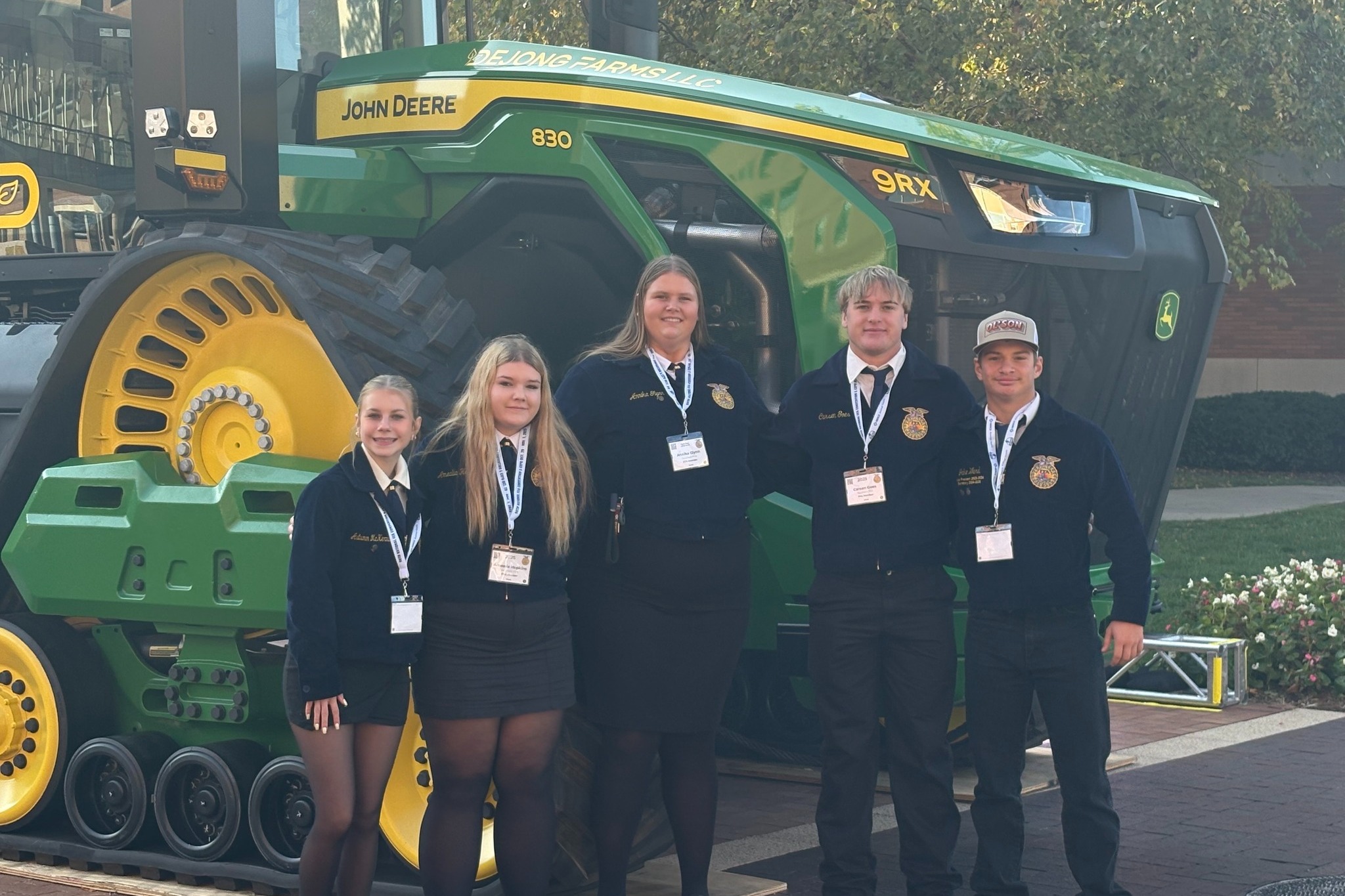 A group of five FFA students stand together in front of a large green John Deere tractor. They are dressed in official FFA jackets and name badges, smiling for the photo during an outdoor event.