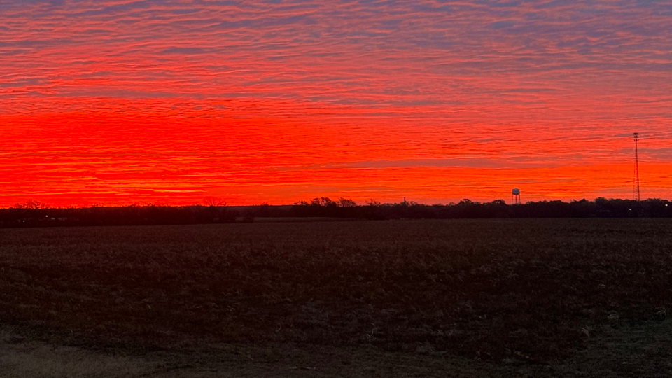 A vivid red and orange sunrise spreads across the sky over a wide rural landscape, with silhouetted trees, a water tower, and a tall communication tower visible on the horizon.