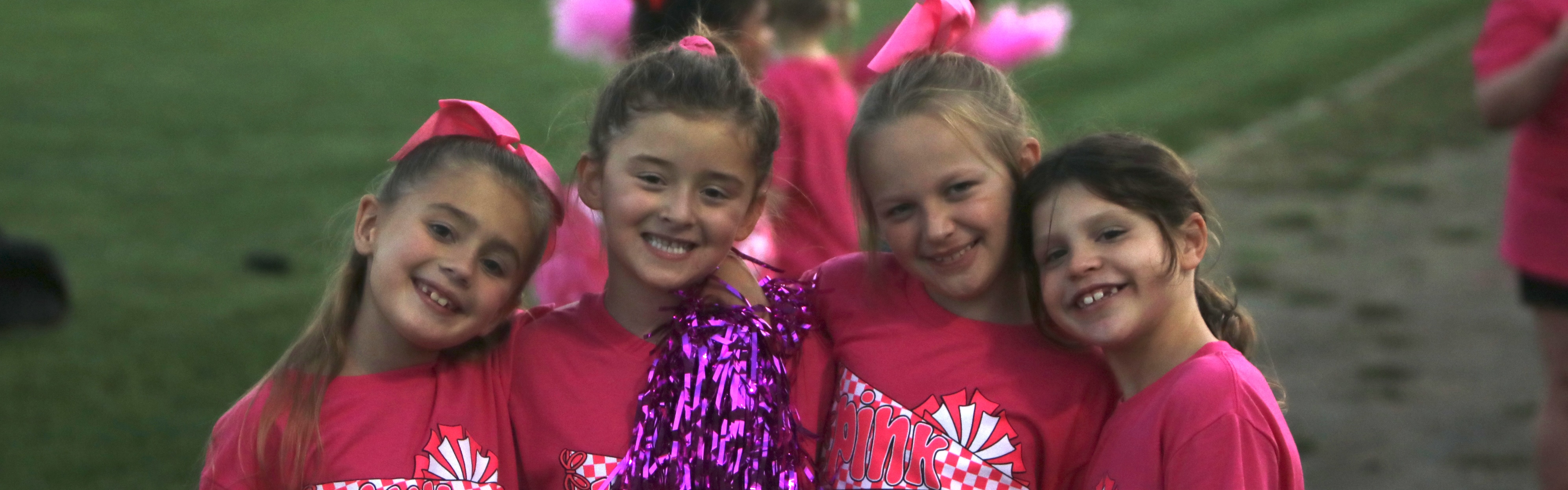 Four young girls wearing matching pink “Pink Out” shirts and hair bows smile and pose together on the sidelines of a field. Two of them hold shiny purple pom-poms, and other children in pink can be seen in the background.