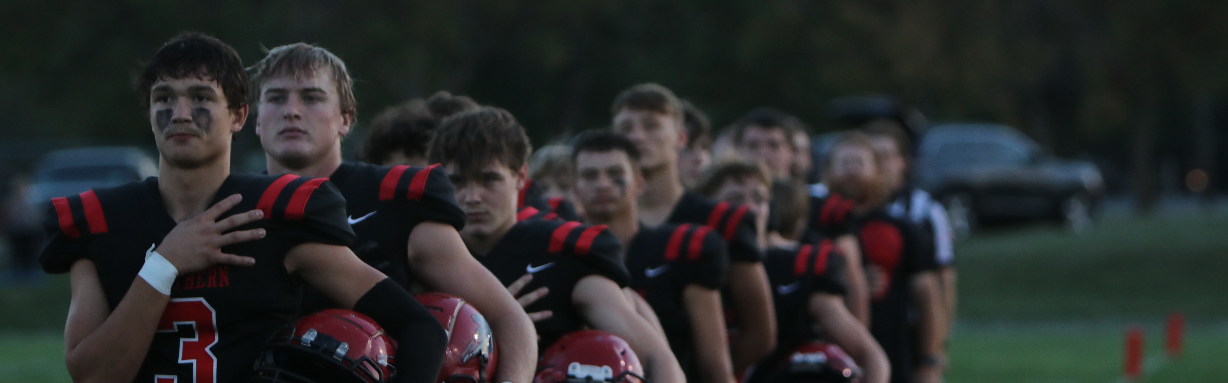 A line of Southern football players stands on the field before a game, each with one hand over their heart and their helmets tucked under the other arm. The player in front wears jersey number 3, and the team is dressed in black uniforms with red accents. Trees and parked cars are visible in the background as dusk begins to fall.