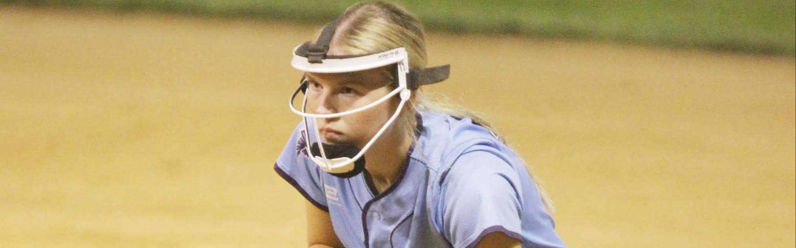 A Freeman-Southern-Lewiston softball player wearing a light blue jersey, navy pants, and protective face mask crouches in a ready position with her glove extended toward the dirt infield during a nighttime game.