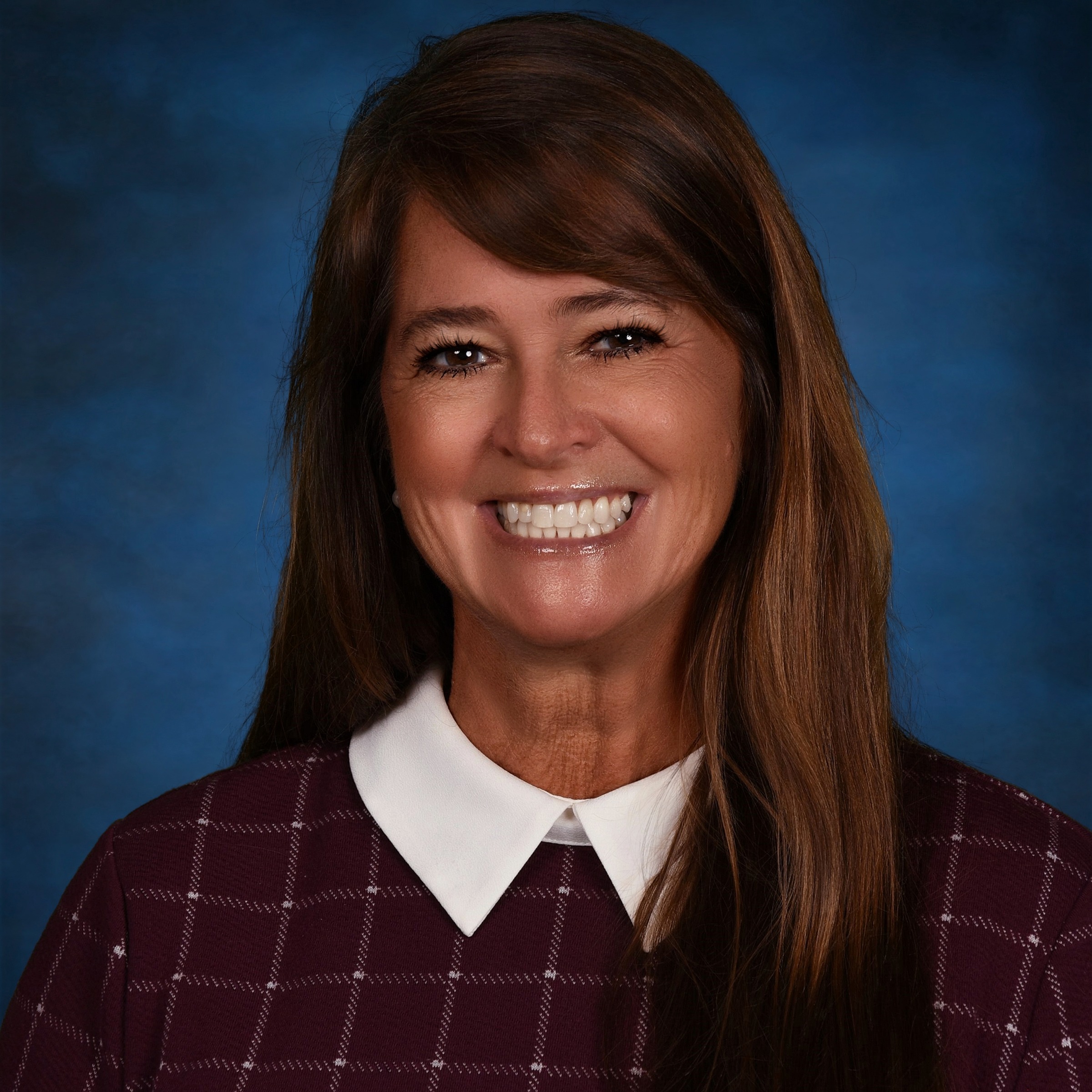 A headshot of a woman with brown hair and a burgundy sweater with a white collar, in front of a blue background.