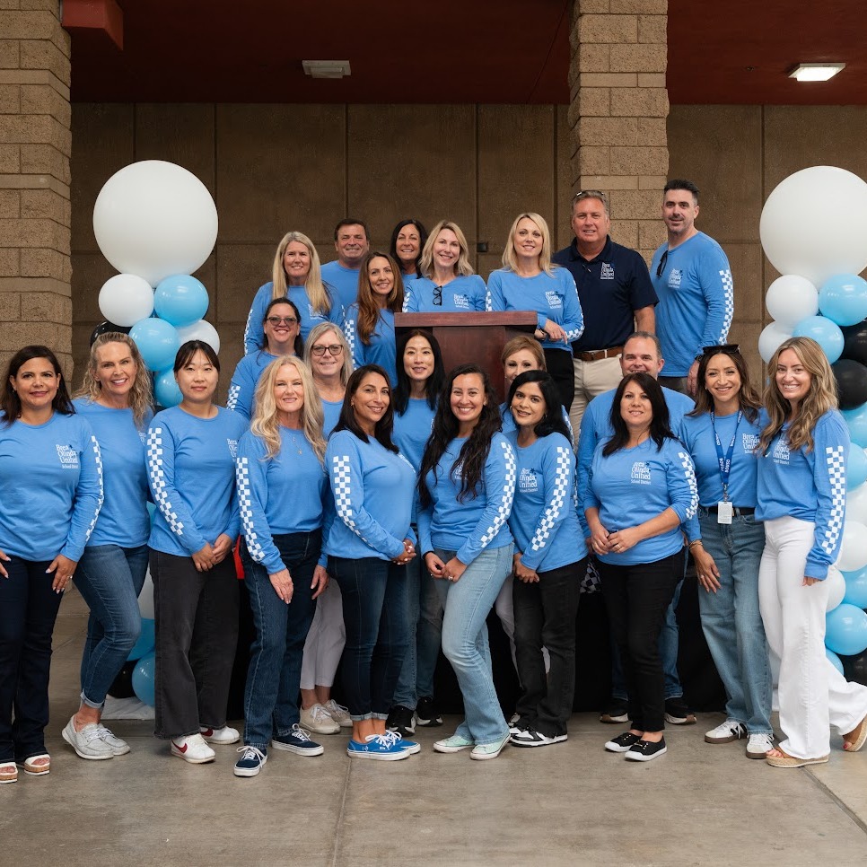 A group photo of members of the district office. employees are wearing blue shirts and are standing in front of white balloons