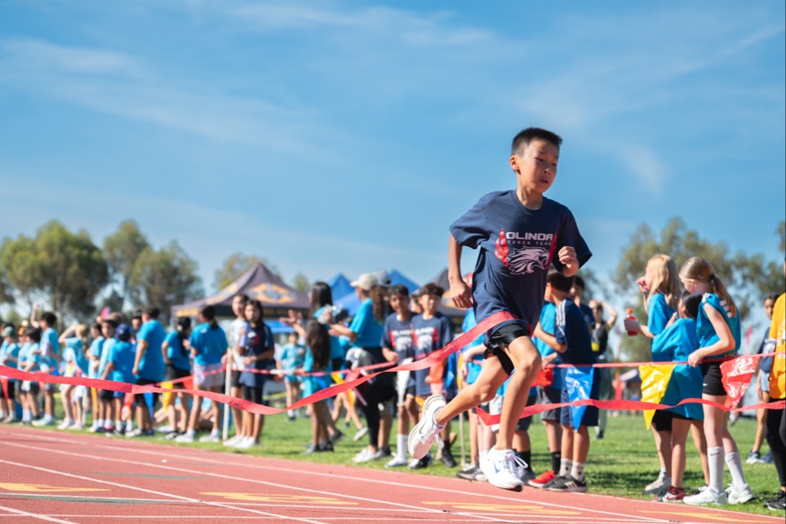 a boy runner crosses the ribbon on a track field. a crowd of other students look on
