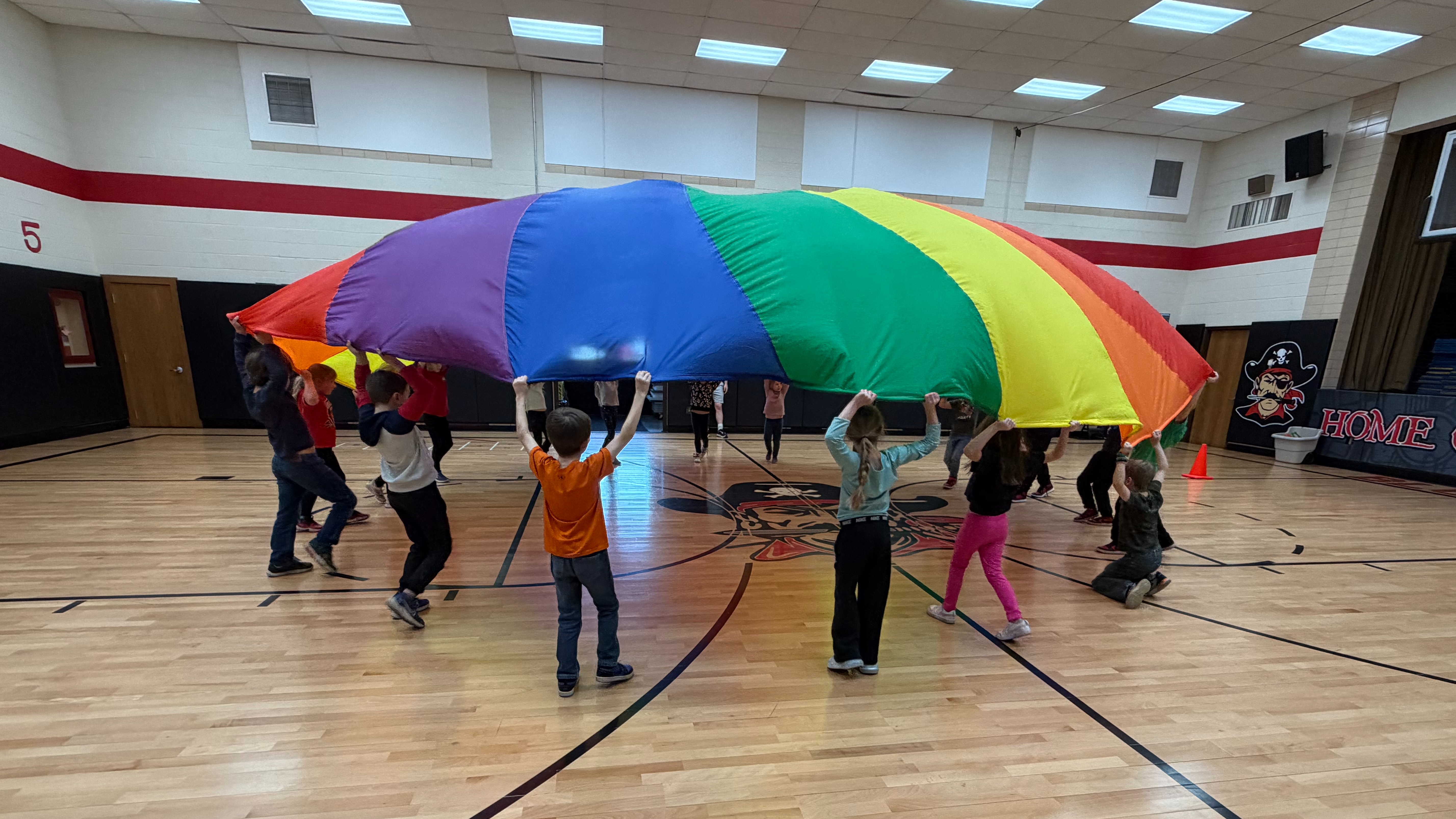 A group of children hold a colorful parachute on a gym floor, with a basketball court layout.